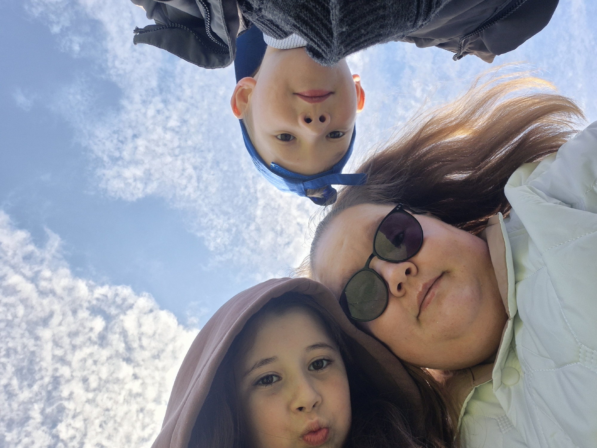An outdoor, low-angle, upward-facing shot captures three individuals against a bright blue sky with scattered white clouds. The perspective suggests the photo was taken from below, with the subjects looking down at the camera.
At the top center, a young boy with light hair wears a blue baseball cap and a dark jacket or sweater. He looks directly at the camera with a subtle smile. To his right, partially obscuring the top right corner, is an adult woman. She wears dark sunglasses and a light-colored jacket and also looks towards the camera with a neutral expression. In the bottom left, a young girl with dark hair is visible, wearing a light brown hoodie. She looks towards the camera with her lips pursed in a pout.
The clear sky with distinct white clouds indicates it is daytime with fair weather. The individuals appear to be posing for a casual photograph, possibly a selfie. No visible text is present in the image. The scene is set in Chaikovsky, Russia.
