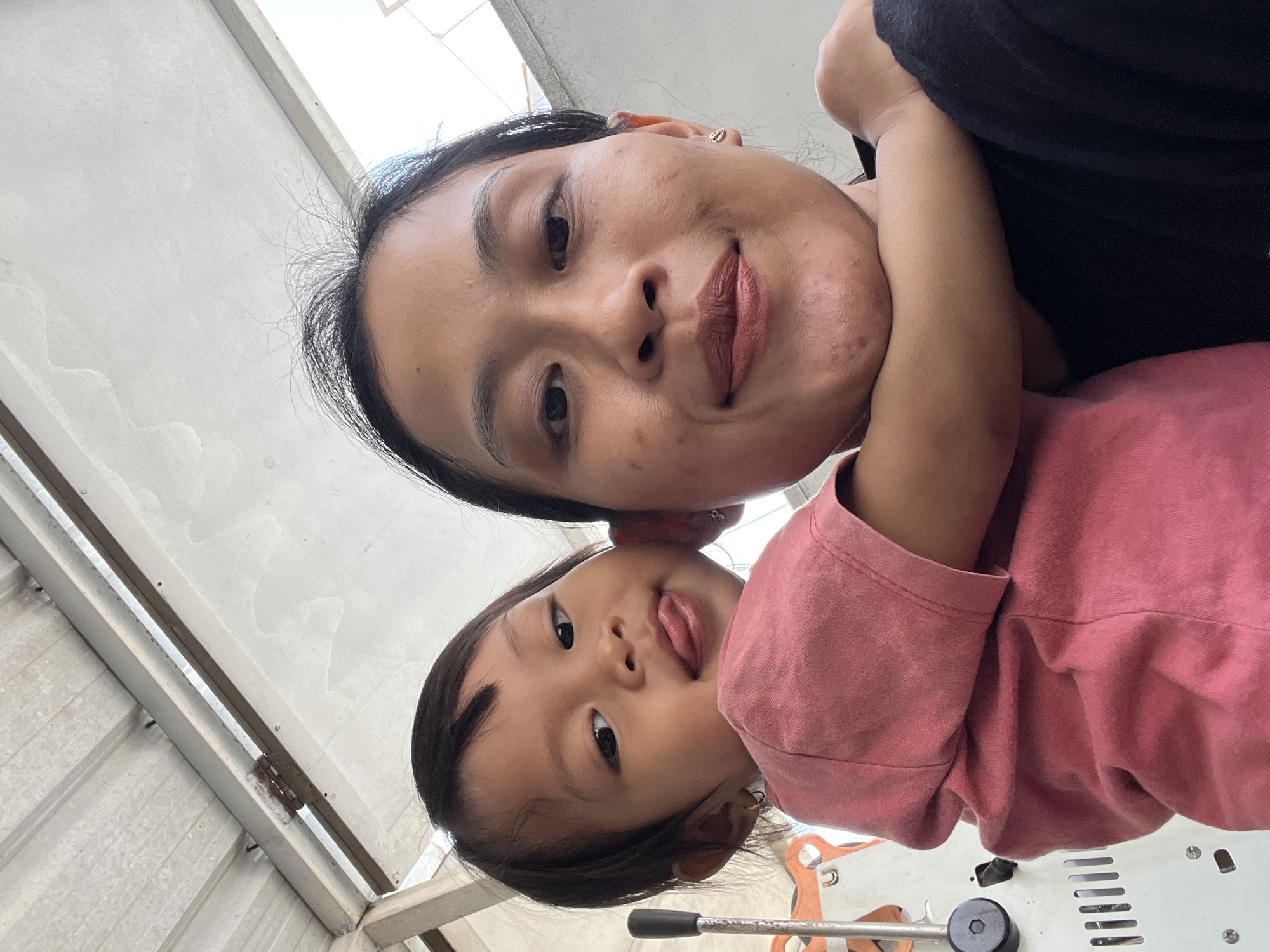 A close-up, upward-angled selfie shows two individuals, an adult female and a young child, positioned centrally. The adult, facing the camera, has dark hair pulled back and is wearing makeup. Her right arm is around the child. The child, leaning in from the left, is wearing a pink t-shirt and has their tongue sticking out. The background is an overhead view of a corrugated metal roof structure, suggestive of an outdoor covered area. The lighting indicates it is daytime. The image does not contain any specific visual cues that definitively place it in Kecamatan Pati, Indonesia.