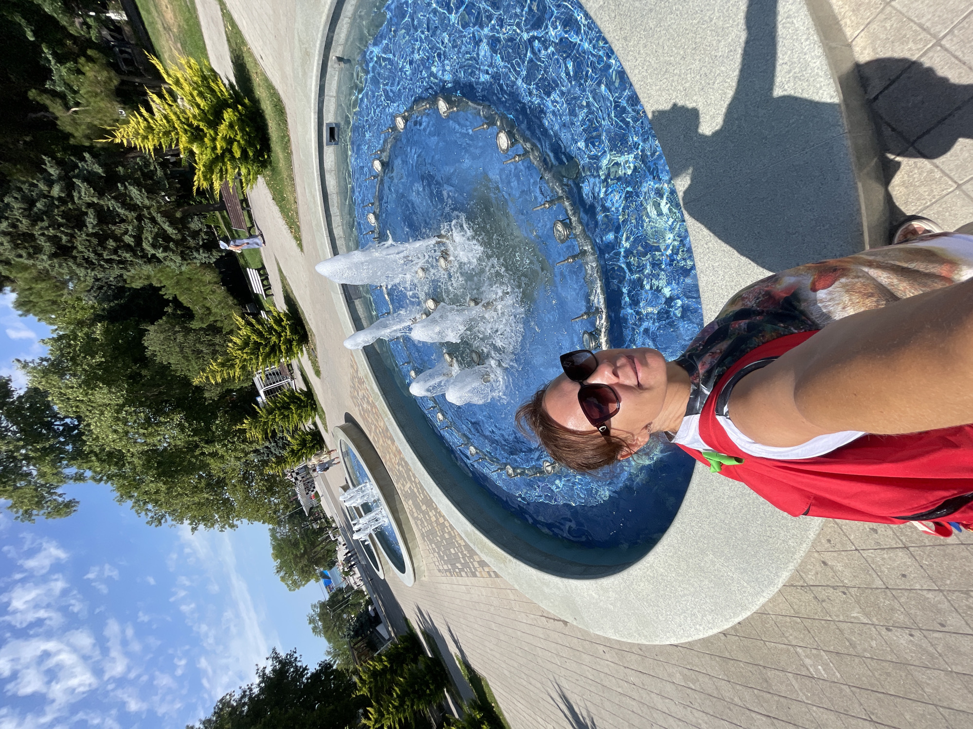 A woman with brown hair and sunglasses takes a selfie in front of a circular fountain in Anapa, Russia. She is wearing a white shirt and a red backpack. The fountain has bright blue water and jets of water shooting into the air. The background shows a park with trees, benches, and a paved walkway. The sky is blue with some white clouds. In the distance, there are more fountains and people walking. The overall scene depicts a sunny day in a public park.