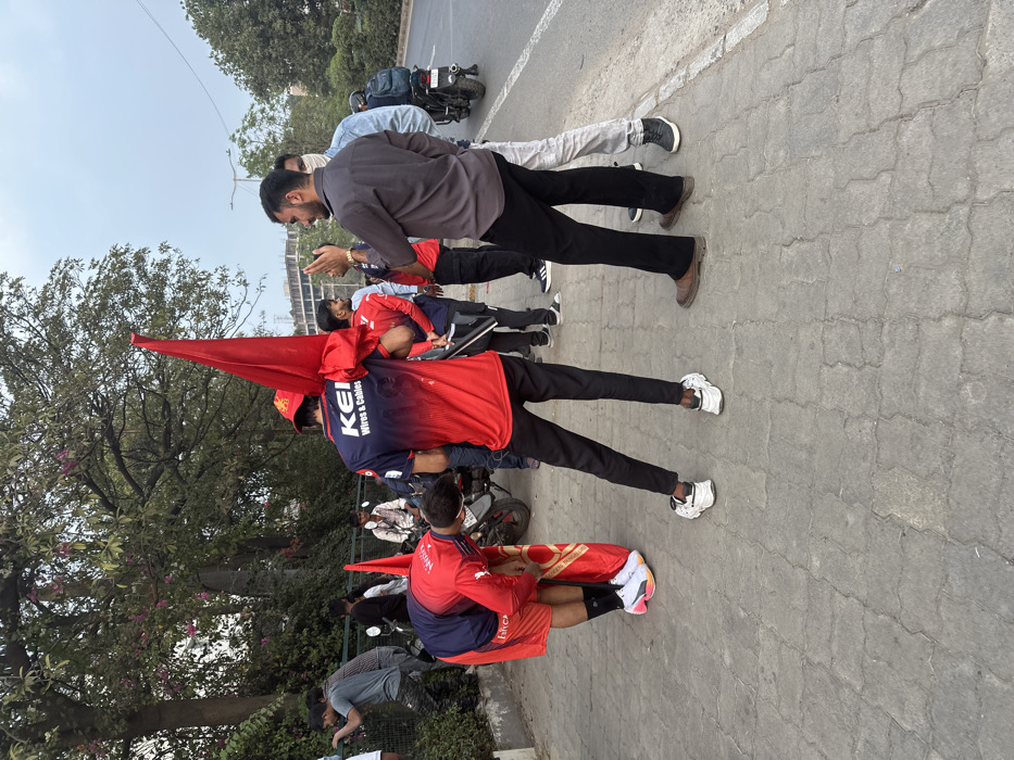 An outdoor scene in Vadodara, India, depicts a group of individuals on a paved sidewalk adjacent to an asphalt road. In the foreground, two male subjects are prominent. One subject, wearing a red and blue shirt with "KEI Wires & Cables" text, red shorts, and white and multicolored sneakers, is bent forward, adjusting a red and gold banner/flag. The banner displays what appears to be a "Vadodara" logo. Immediately behind him, another male subject, also wearing a red and blue "KEI Wires & Cables" shirt, black pants, and white sneakers, stands with his back to the viewer, holding a red flag upright over his left shoulder.
To the right, a male subject in a dark grey button-up shirt and black pants is gesturing with both hands while facing left. Next to him, a male subject in a light blue shirt and light-colored pants wears a light-colored face mask. Further right, a motorcycle is parked on the roadside, with another male subject on it, wearing a blue backpack.
On the left side of the frame, several other individuals are visible near a green metal fence and dense foliage. One male subject in a grey t-shirt and camouflage-patterned pants is bent over, seemingly looking through the fence. Trees with sparse foliage and hints of pink blossoms are visible above the fence. The background features a partly cloudy sky and distant urban structures, including a building under construction. The sidewalk is paved with grey interlocking blocks.