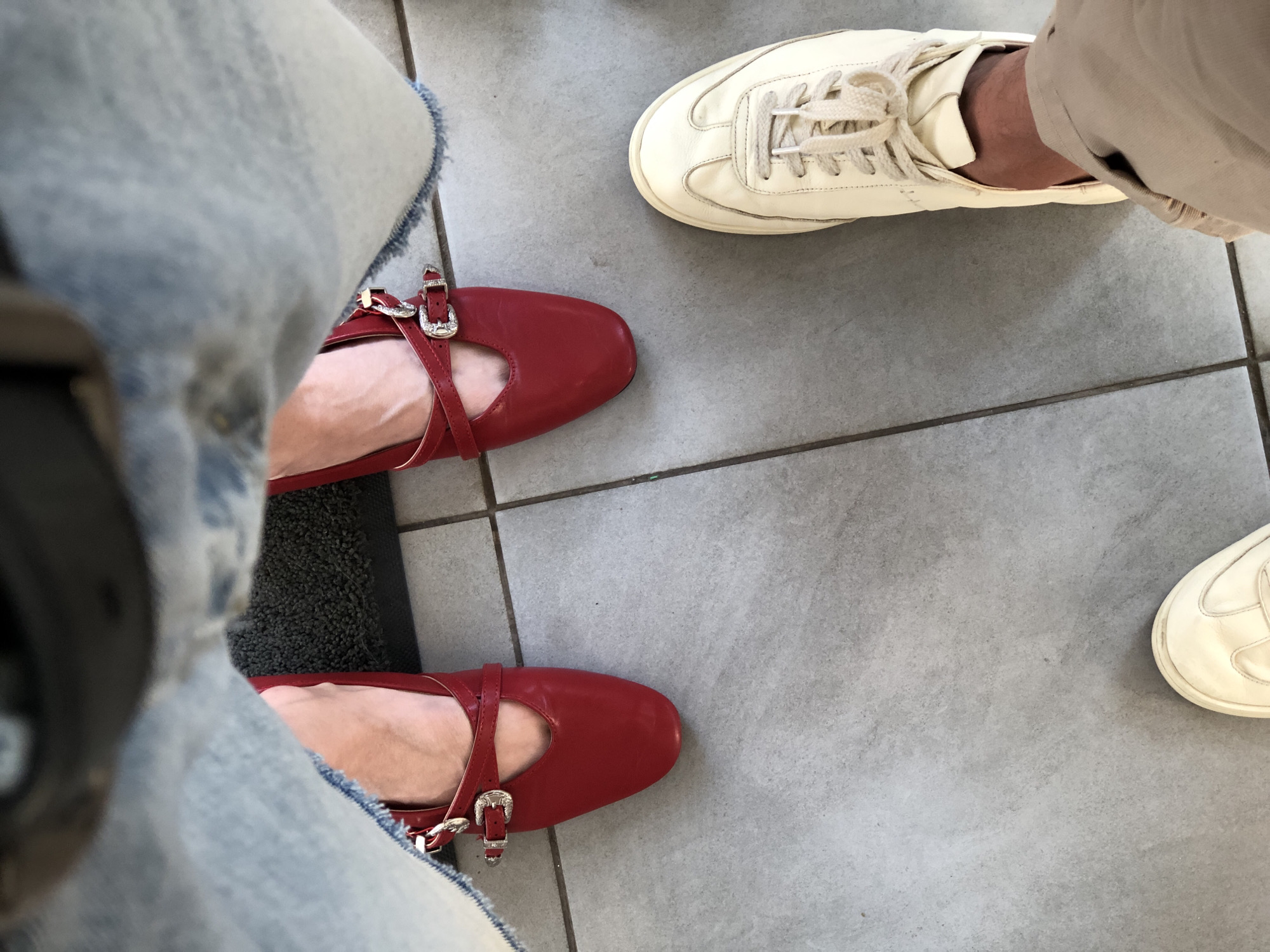 The image shows a low-angle view of two people's feet, standing on a tiled floor. One person is wearing red ballet flats with buckle straps, and the other is wearing off-white sneakers. The tiled floor is light gray with darker grout lines. In the background, part of a dark mat is visible, and above that, the denim of jeans is visible. The scene appears to be indoors, possibly in a public space or a home. There is no specific indication of the location being Saint Petersburg, Russia.