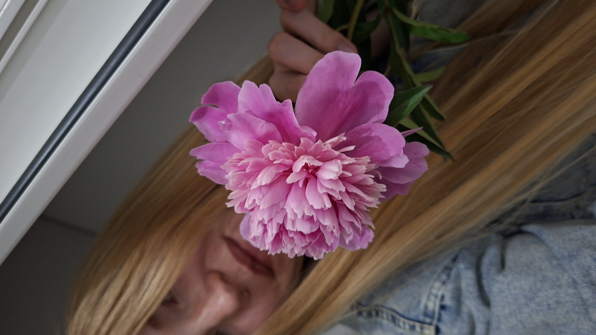 An overhead, close-up shot shows a person wearing a denim jacket, holding a pink peony. The person's face is out of focus in the background, but their long blonde hair is visible framing the flower. The peony is the central subject of the image, with its delicate petals in sharp focus. The background is blurry, showing a light-colored wall and a dark surface. The image appears to have been taken indoors. No specific location in Chaikovsky, Russia, can be determined from the visual information provided.