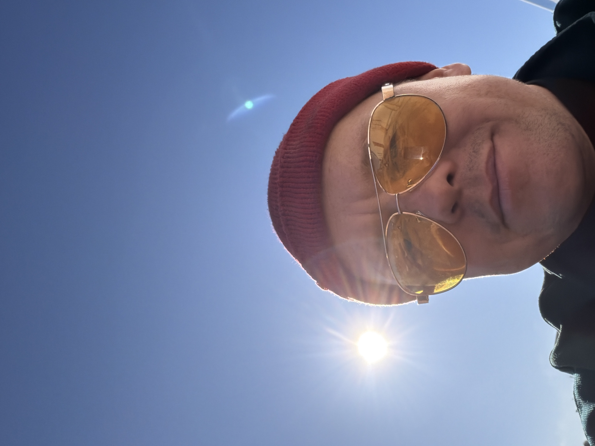 A low-angle, upward-facing selfie captures a person wearing a red knit hat and gold-rimmed aviator sunglasses. The person is positioned in the upper right quadrant of the image, partially obscuring the clear blue sky, where the sun is shining brightly near the bottom of the frame. The person's face is partially visible, showing part of the forehead, nose, and mouth. A portion of a dark-colored garment is visible on the right side of the frame. The location is outdoors, with the clear sky and bright sunlight indicating daytime. There are no indicators of the location being in the city of Chaikovsky, Russia.