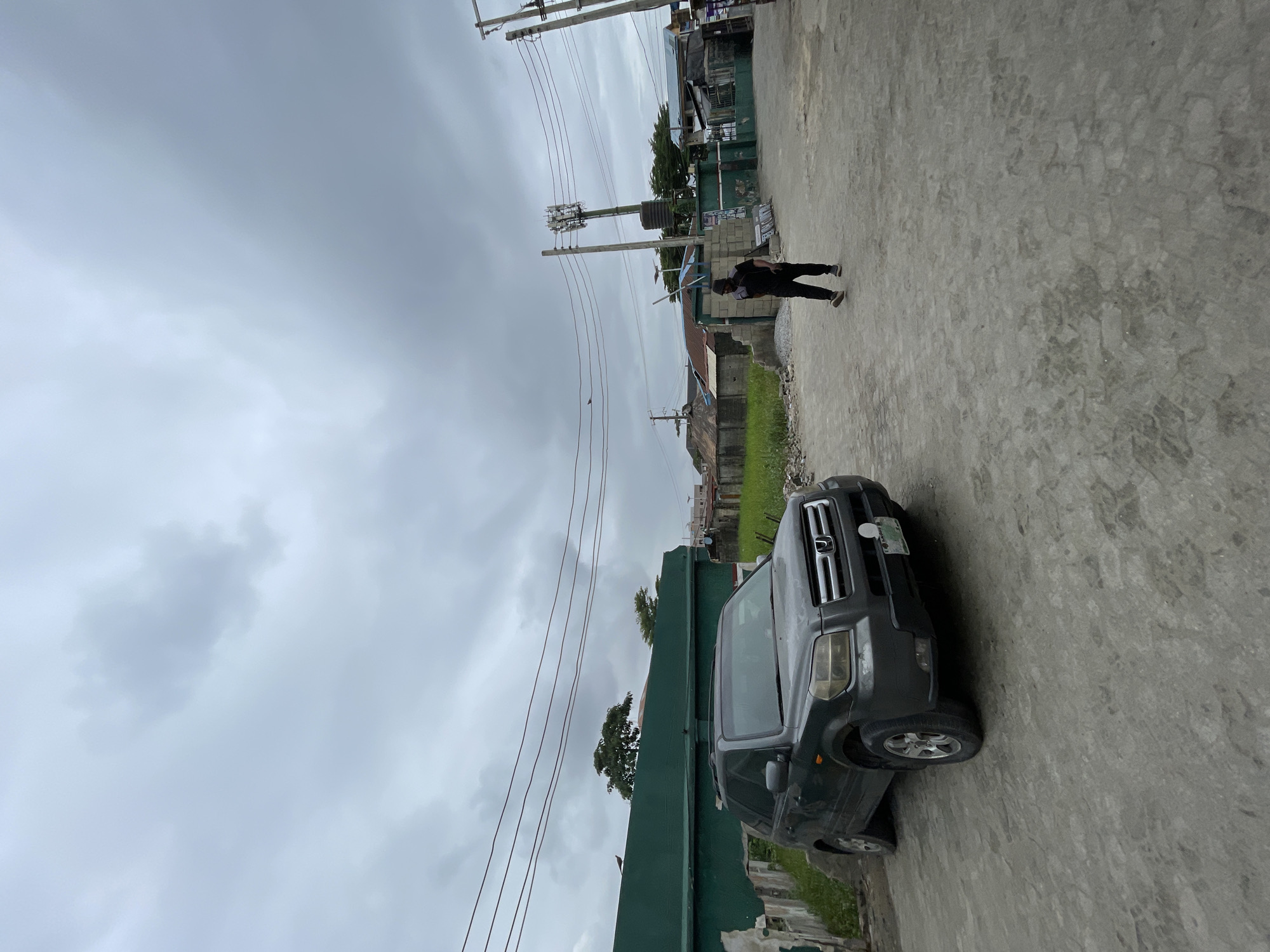 In Ado, Nigeria, a dark gray Honda Pilot SUV is parked on a dirt and gravel lot under an overcast sky. The vehicle faces left, with its front bumper and driver's side visible. Behind the car, a low green wall runs horizontally across the frame. In the middle ground, a man dressed in dark clothing walks away from the camera towards the right, his gait suggesting movement. To his right, a tall cell tower with multiple antennas rises above other structures. The background features a mix of residential buildings with corrugated metal roofs and some vegetation. Power lines crisscross the sky, and the overall environment appears to be an urban or semi-urban setting.