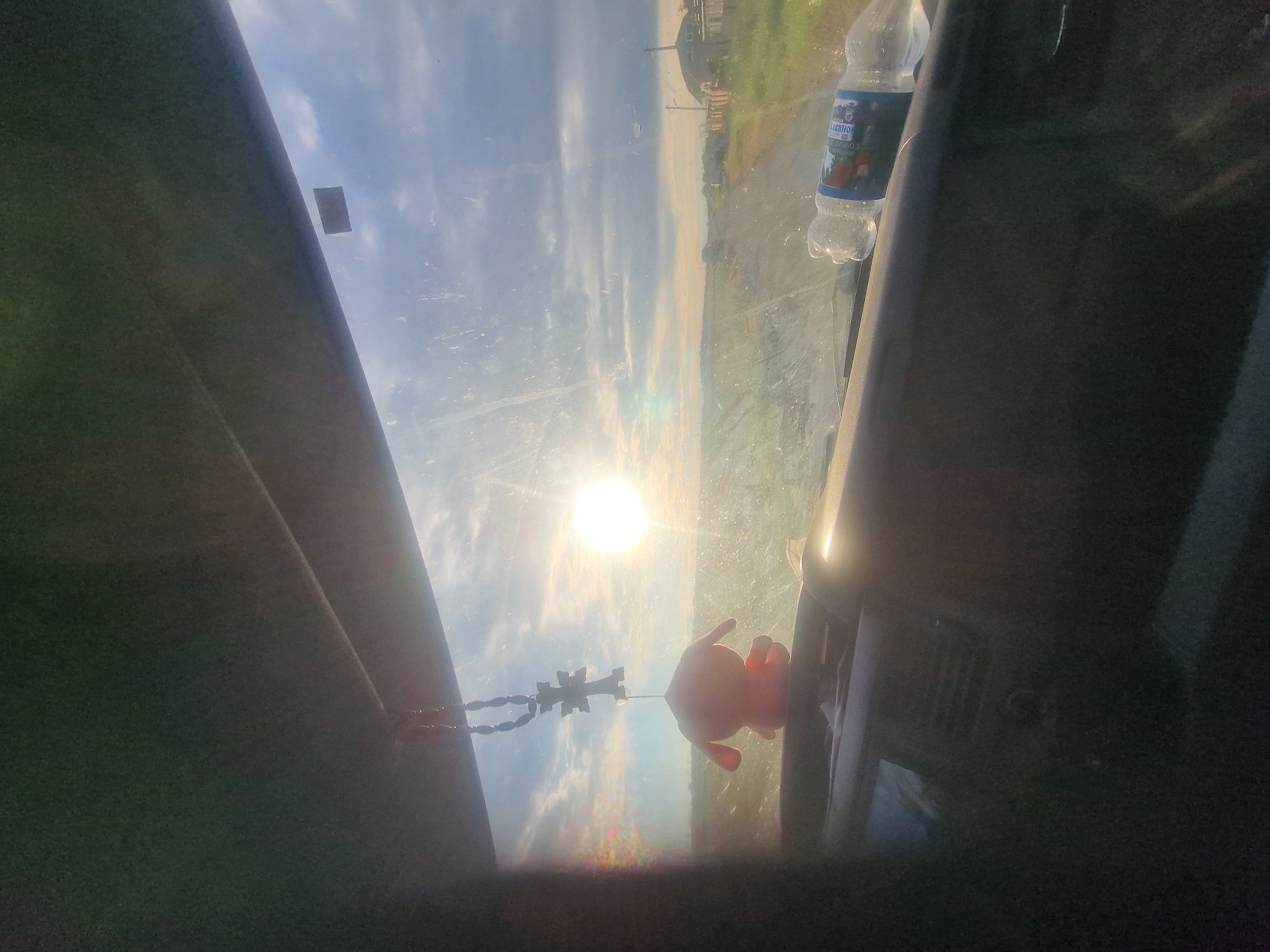 The image is taken from inside a vehicle, looking through the windshield at a rural landscape in Kursavka, Russia. The sun is setting, casting a warm glow and creating lens flare across the scene. In the foreground, on the dashboard, there is a small, pink, stuffed animal and a cross-shaped pendant hanging from the rearview mirror. A partially consumed bottle of water is also visible on the dashboard. The road ahead is unpaved and leads into the distance, with green fields and scattered houses on either side. The sky is partly cloudy, with dramatic cloud formations illuminated by the setting sun.
