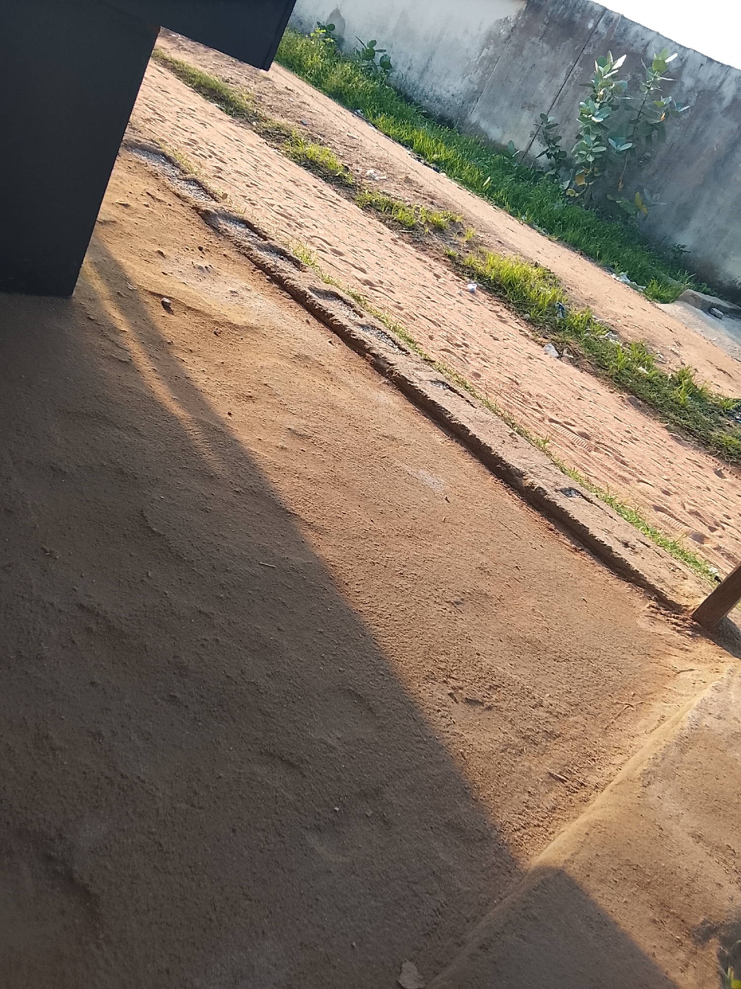 This image shows an outdoor scene in Jalingo, Nigeria. The foreground is dominated by a dusty, sandy ground, with a distinct shadow cast across it, suggesting it is likely morning or late afternoon. A portion of a large, dark, rectangular object, possibly furniture or part of a structure, is visible on the left side of the frame.
The ground slopes upwards away from the viewer, meeting a concrete wall in the background. A narrow strip of green grass and some small plants grow along the base of the wall. A few scattered pieces of debris are visible on the sandy surface. The overall impression is of a somewhat neglected outdoor area, perhaps a yard or a pathway. The lighting indicates clear weather. There is no visible text in the image.