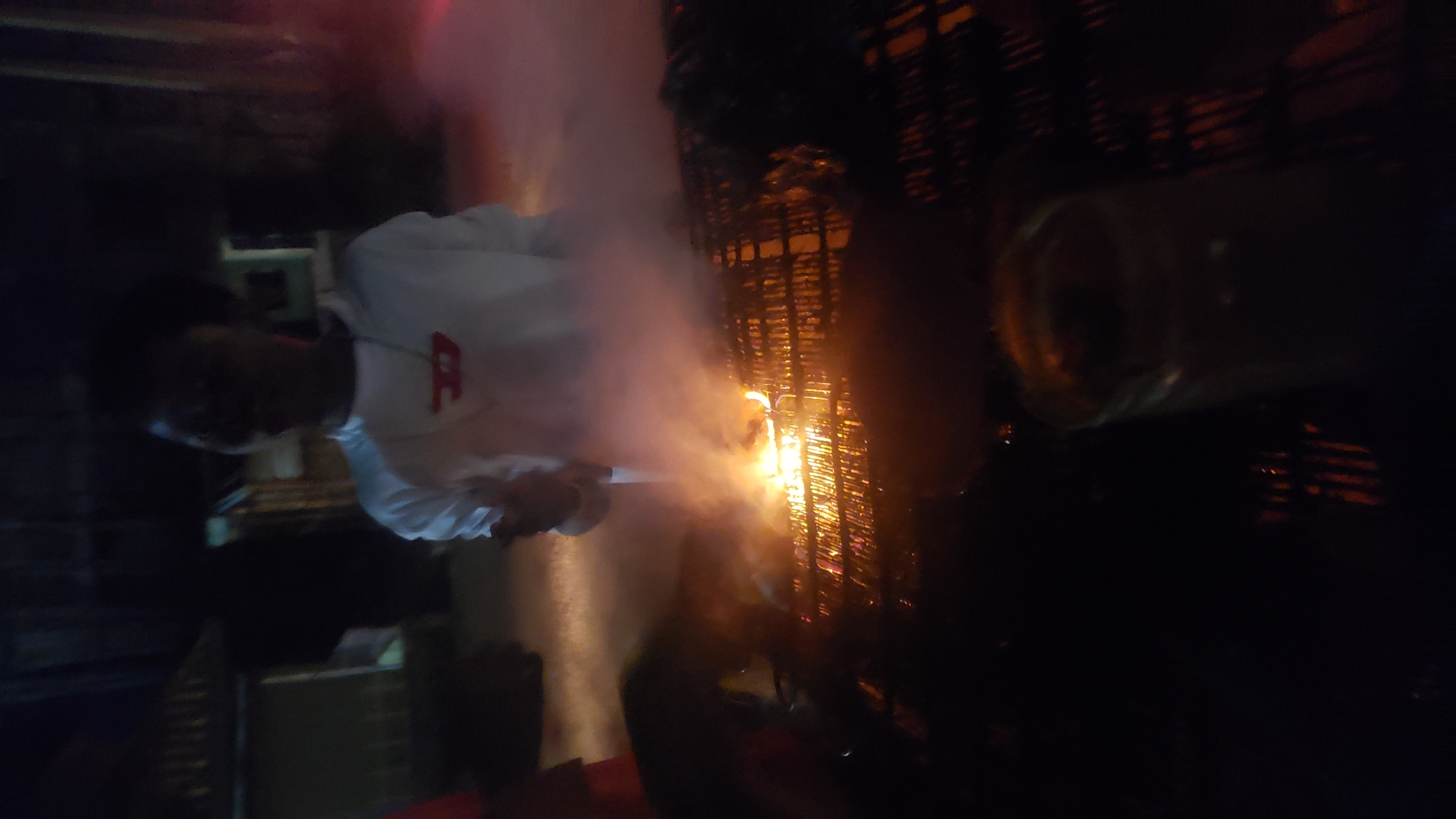 This nighttime image captures an outdoor scene in Narayi, Nigeria. A male individual, approximately centered in the upper frame, is visible wearing a white long-sleeved shirt with a red logo on the chest and a visible necklace. His face is indistinct due to low light and blur. In the middle ground, a metal barbecue grill contains active flames and dense smoke emanating primarily from its left side. A blurred glass or bottle containing a beverage is present in the bottom right corner of the frame. The background is dark, revealing faint structural outlines consistent with buildings or shelters. The primary light source originates from the burning embers and flames on the grill.