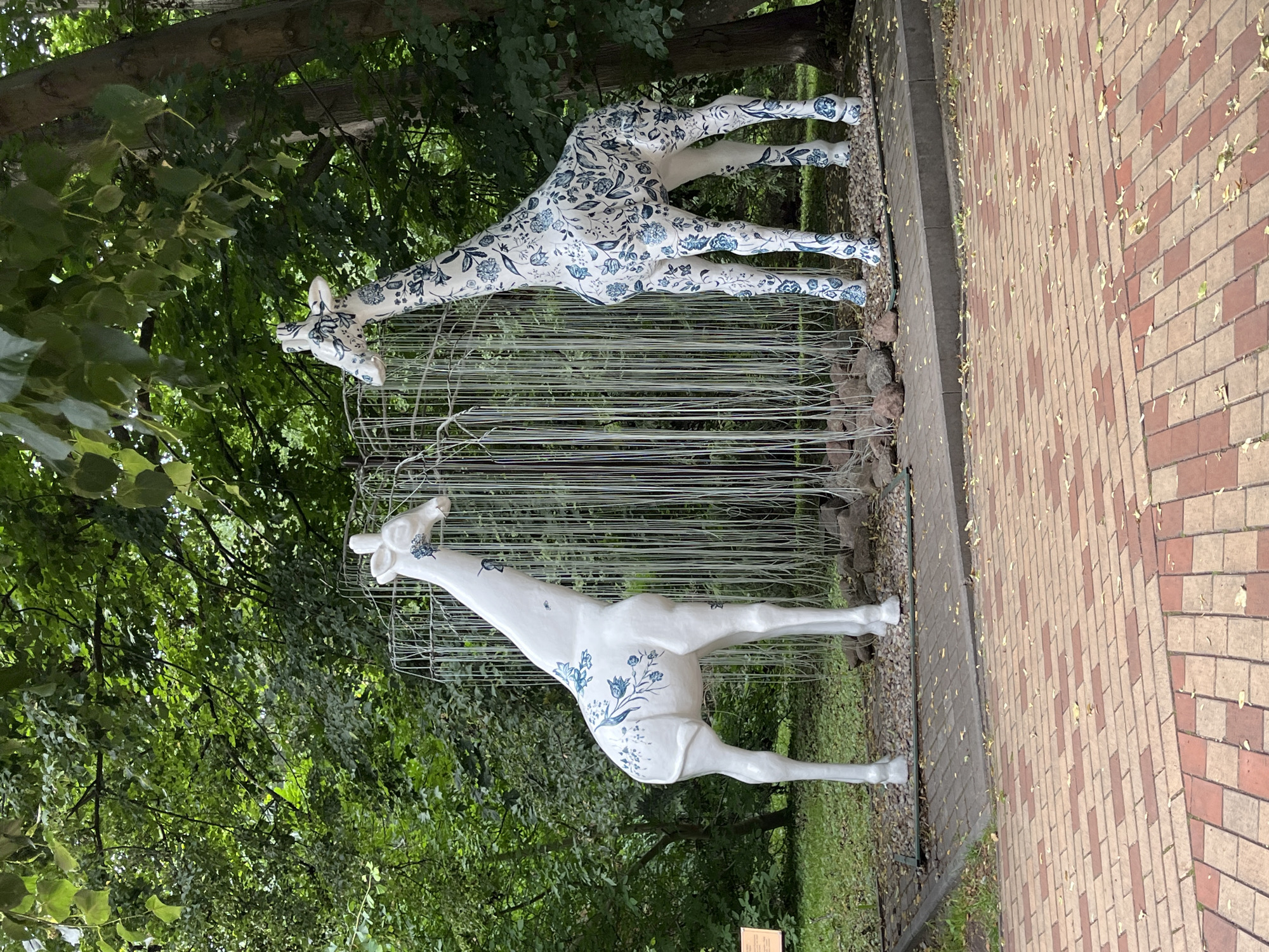 Two white giraffe sculptures, decorated with blue floral patterns, stand in a park in Yantarny, Russia. The sculpture on the left is taller and more slender than the one on the right. Both giraffes are facing towards each other, with a decorative structure resembling a weeping willow tree behind them. The ground in front of the sculptures is paved with reddish-brown and beige bricks, and scattered leaves are visible. The background consists of lush green trees and foliage, with a thick tree trunk to the right of the image. A small sign is visible on the left side of the image, near the giraffe. The scene is outdoors and appears to be daytime.