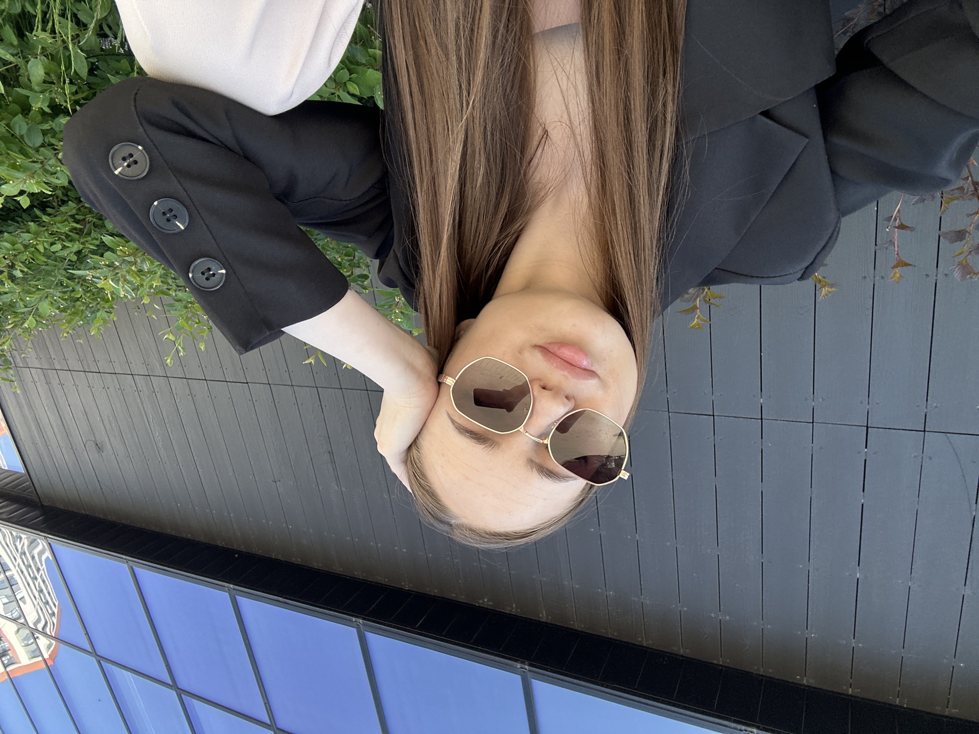 A female individual is depicted, wearing sunglasses and a black blazer with buttoned sleeves. She is positioned near a modern building with blue glass panels, likely reflecting the sky or nearby structures. The location appears to be an outdoor space in a city setting, with some greenery present. The photo is taken at an unusual angle.