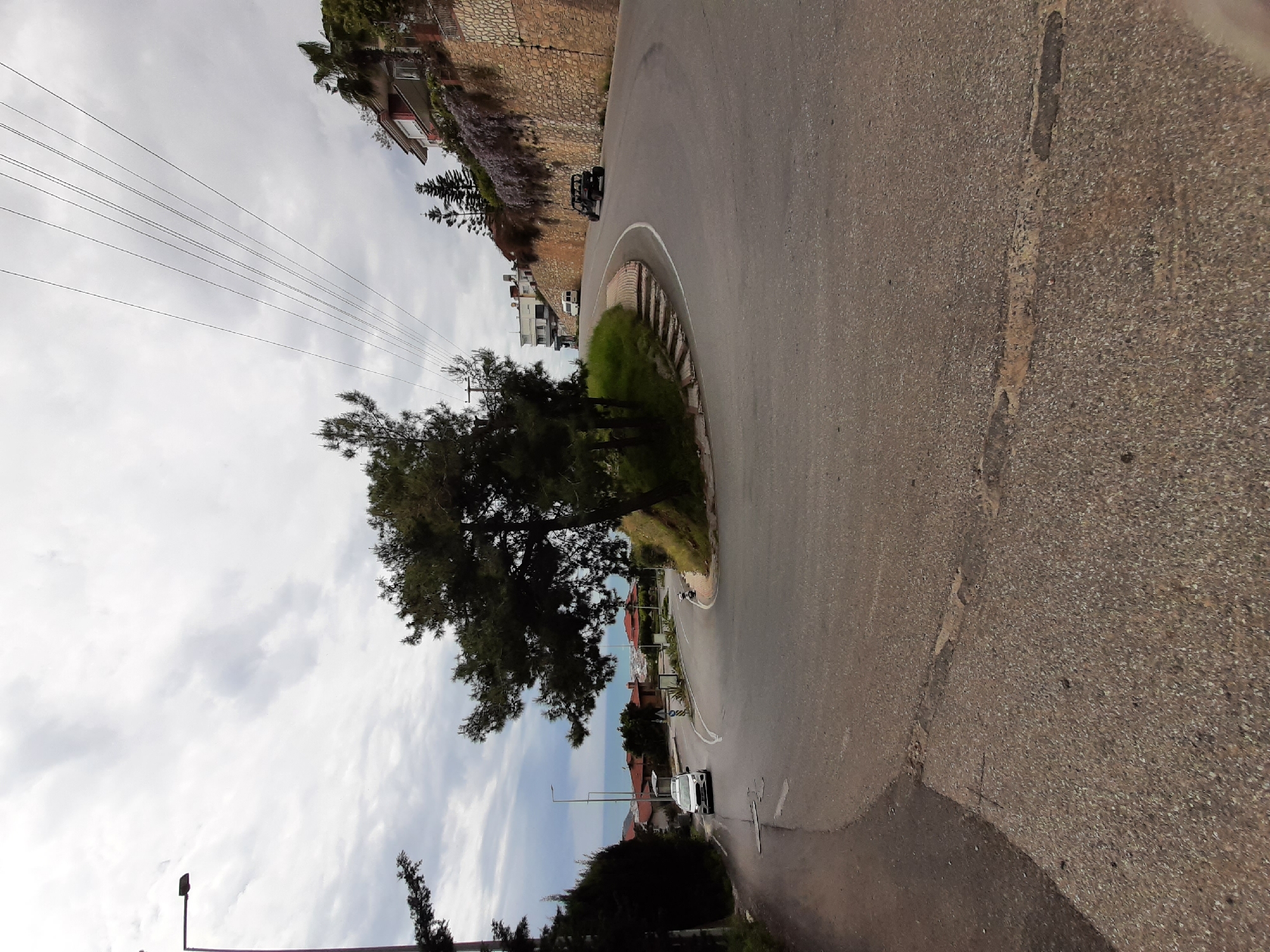 The image depicts a winding road in Alanya, Turkey. The road curves sharply to the right, with a concrete barrier and vegetation on the inside of the curve. The road surface is asphalt, showing some wear and tear. There are several cars and motorcycles on the road. In the background, there are buildings and trees, with the sky partially visible through the branches. Power lines are visible overhead. The overall scene suggests a residential area located on a hillside.