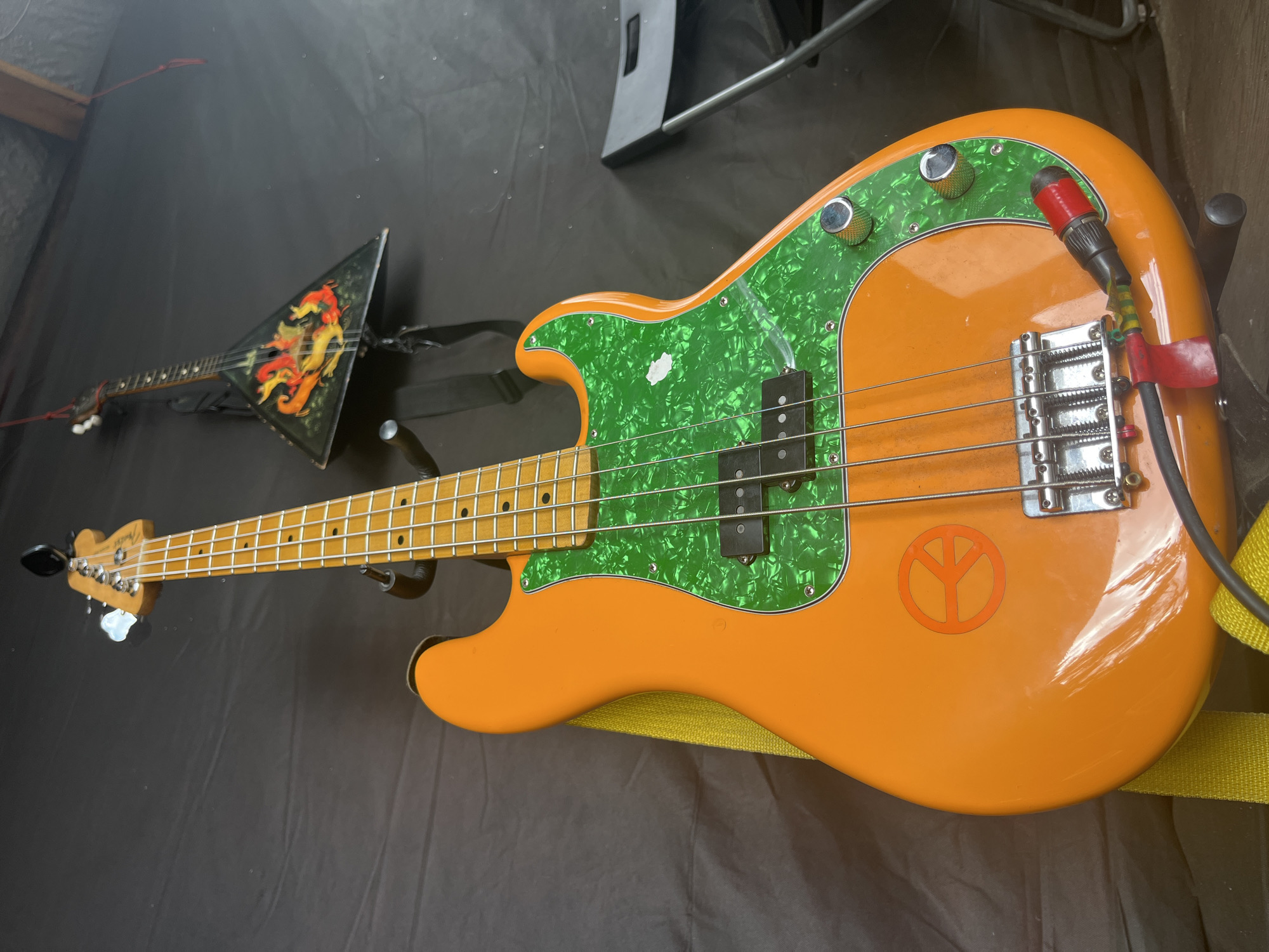 In an indoor setting with a dark fabric backdrop, an orange bass guitar is prominently displayed. The guitar features a green, pearlescent pickguard and a peace sign decal on its body. A yellow strap is visible, attached to the guitar. To the left of the bass guitar, a black chair is partially visible. In the upper background, a triangular stringed instrument, possibly a balalaika, is hanging and decorated with a fiery design. The floor appears to be made of wood. The scene is static and there are no people present.