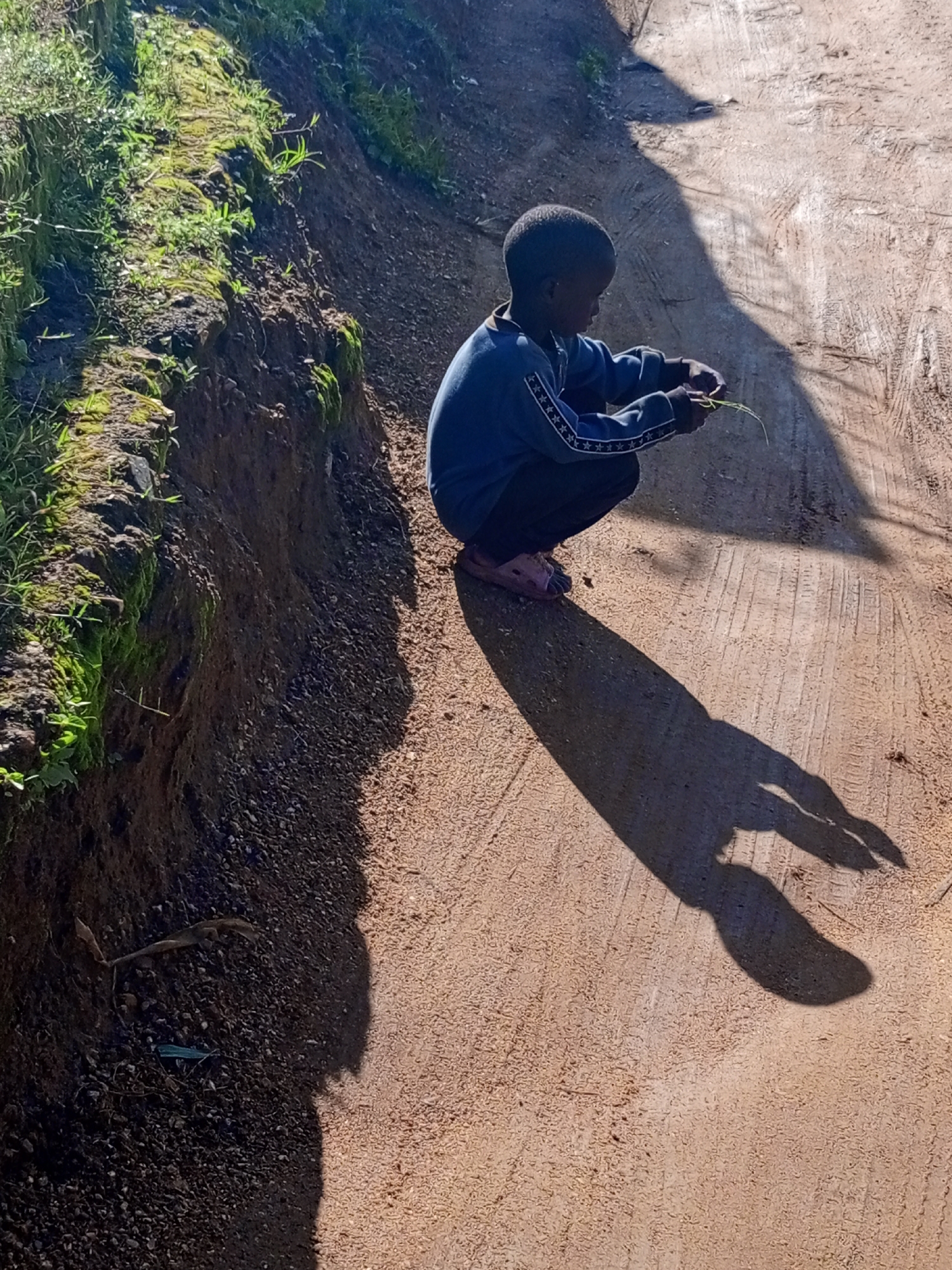 A young person, with dark skin and short dark hair, is crouched on a dirt road, facing to the right and away from the camera. They are wearing a dark blue sweatshirt with a white star pattern along the sleeves and dark pants, and pinkish-brown sandals. Their hands are in their lap, and they appear to be holding a thin green blade of grass. A long shadow stretches out from the person across the road, suggesting the sun is to their left and behind them.
The road is composed of light brown, sandy dirt with tire tracks visible. To the left of the road, there is a steep, eroded embankment covered with green grass and moss. The embankment is dark brown and appears somewhat damp. Sunlight illuminates the scene, casting strong shadows. The overall environment suggests an outdoor, possibly rural or semi-rural setting in Nigeria, specifically on Rukuba Road.