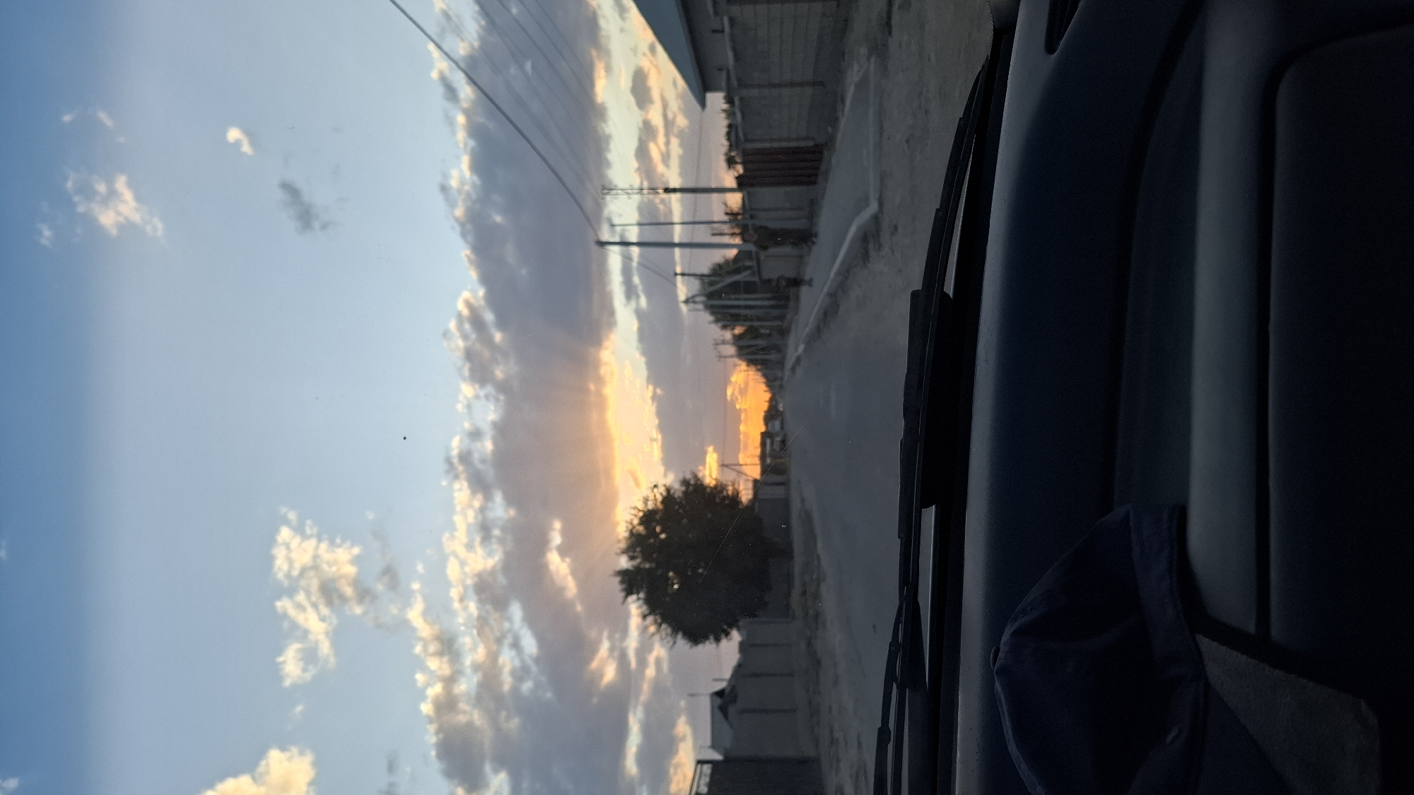 The image, taken from inside a vehicle looking out through the windshield, shows a road in the city of Shubarssu, Kazakhstan at sunset. The sky is a gradient of blue with scattered clouds, some illuminated by the setting sun. The sun itself is low on the horizon, casting rays through the clouds. The road is paved and appears to be in a residential area, with houses and fences visible on either side. A solitary tree stands to the left of the road. Power lines are visible overhead. Two figures, appearing to be children, are walking on the sidewalk on the right side of the road. The dashboard of the vehicle, including a dark-colored cap, is in the foreground.