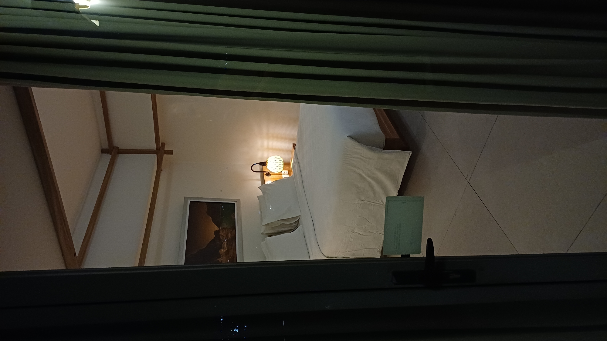 A bedroom scene is viewed through a window or opening obscured by dark green curtains and a window frame. The bedroom is lit by a warm, soft light. A large bed with white linens and pillows is centered in the frame, positioned against a light-colored tiled floor. A bedside table features a lamp with a round shade, emitting a soft glow. A framed picture hangs on the wall behind the bed. The architecture of the room includes exposed wooden beams that form a peaked ceiling. The location is Unknown, Unknown. The window frame appears to include a handle or latch.