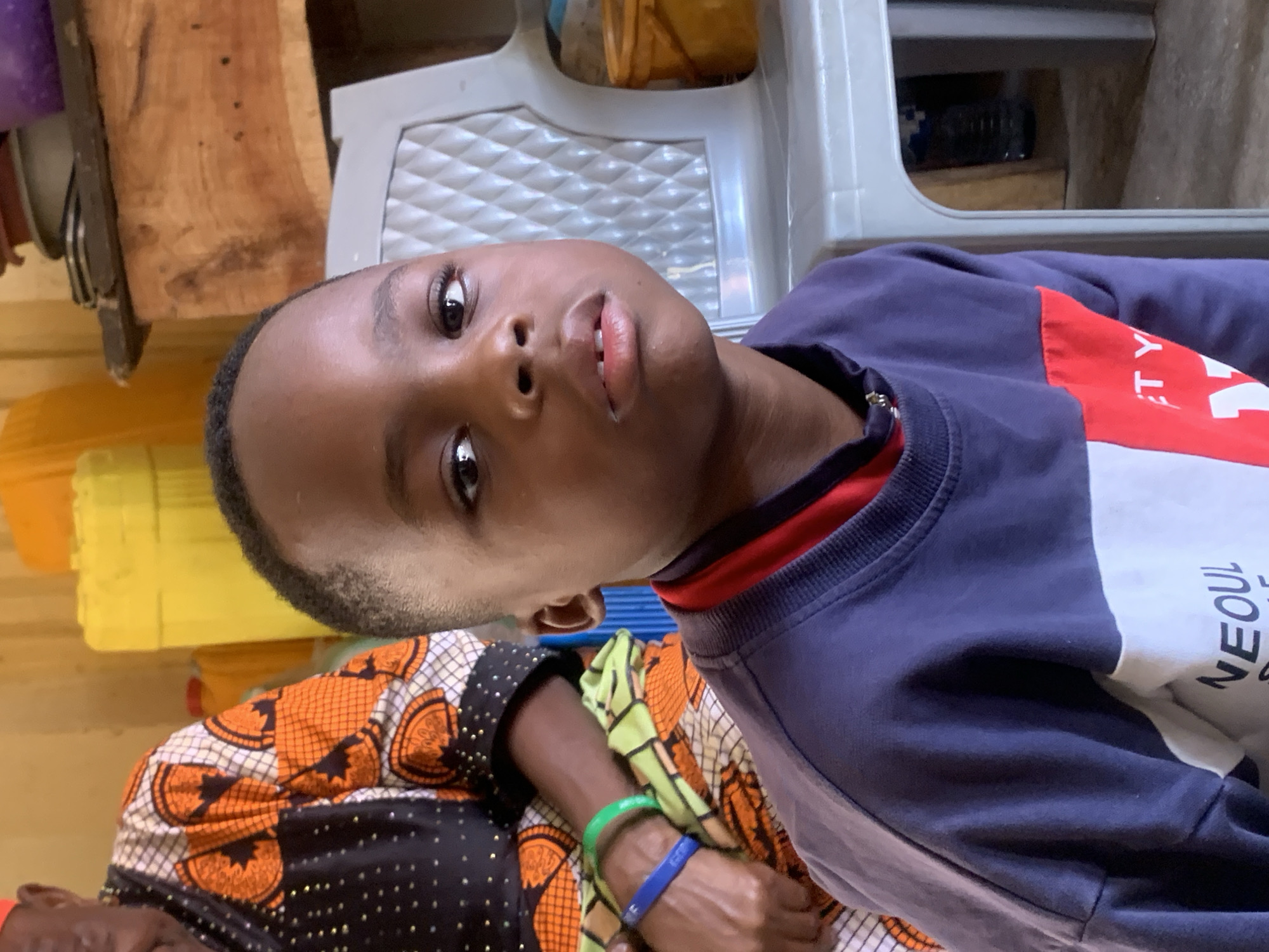 The image depicts a close-up of a young Black male with dark skin and short, dark hair. He is looking directly at the camera with a neutral expression. He is wearing a long-sleeved shirt that appears to be dark blue on the top and white with a red stripe and some text on the lower portion. Behind him, to the left, a portion of an older Black individual is visible, wearing a colorful garment with an orange and black pattern adorned with small white dots and some metallic accents. This individual has bracelets on their wrist, one green and one blue.
The background suggests an indoor environment with a light-colored wall. To the left, there are large yellow containers, possibly water jugs. To the right, a grey plastic chair is prominent, and behind that, a wooden shelf holds several items, including what appear to be potted plants and containers. The floor is visible at the bottom of the frame and appears to be concrete. The scene is lit by natural light.
Based on the provided context, this image was taken in Jos, Nigeria. The presence of large water containers and the general setting are consistent with many residential or communal areas in Nigerian cities. The clothing and physical features of the individuals are also consistent with the demographics of Jos.