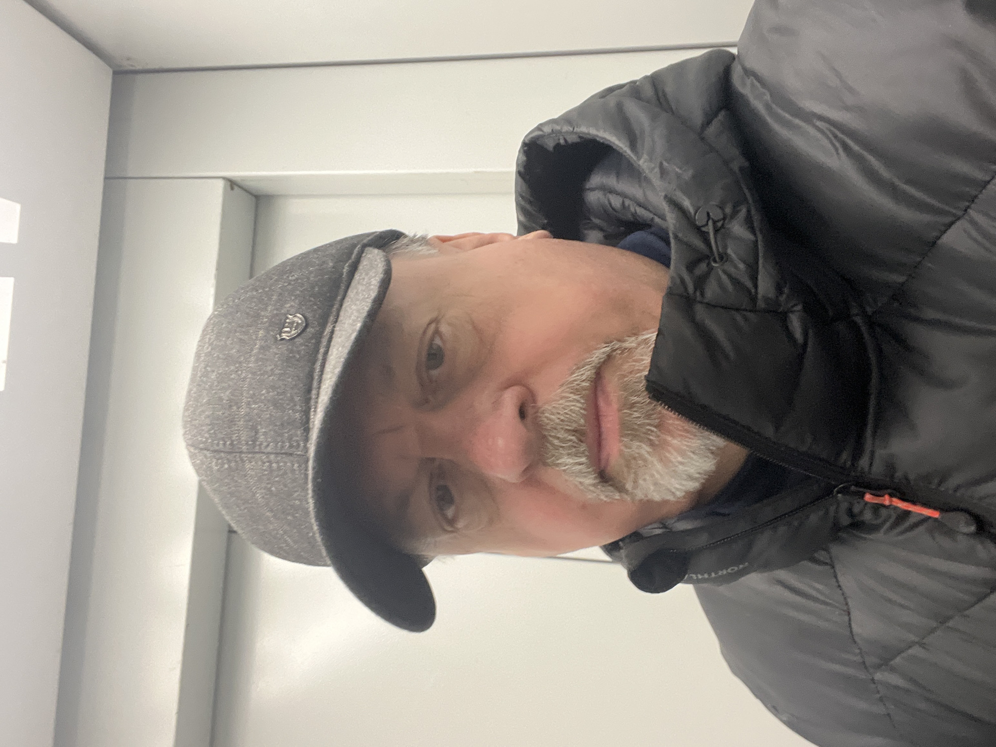 The image depicts a close-up, frontal view of a single male individual positioned indoors. The man appears middle-aged, with a light complexion, dark eyes, and a grey goatee beard. He is wearing a dark grey, textured baseball cap featuring a small metallic shield-shaped emblem on the left front panel. His attire also includes a black quilted puffer jacket with a partially visible "NORTHE" label on the inner collar, suggesting a brand name like "NORTHERN." The jacket has a black zipper with a distinct orange pull tab. A navy blue garment is visible beneath the jacket's collar. The individual is looking directly towards the viewer with a neutral expression, suggesting a selfie or direct portrait.
The immediate environment behind the man consists of plain, light-colored (likely white or very light grey) vertical panels with horizontal seams visible in the upper background. These panels and the enclosed nature of the space suggest the interior of an elevator or a similarly confined, utilitarian room. No specific details within the image context allow for the identification of a precise location within the city of Podolsk, Russia. The lighting is even and consistent with indoor artificial illumination.