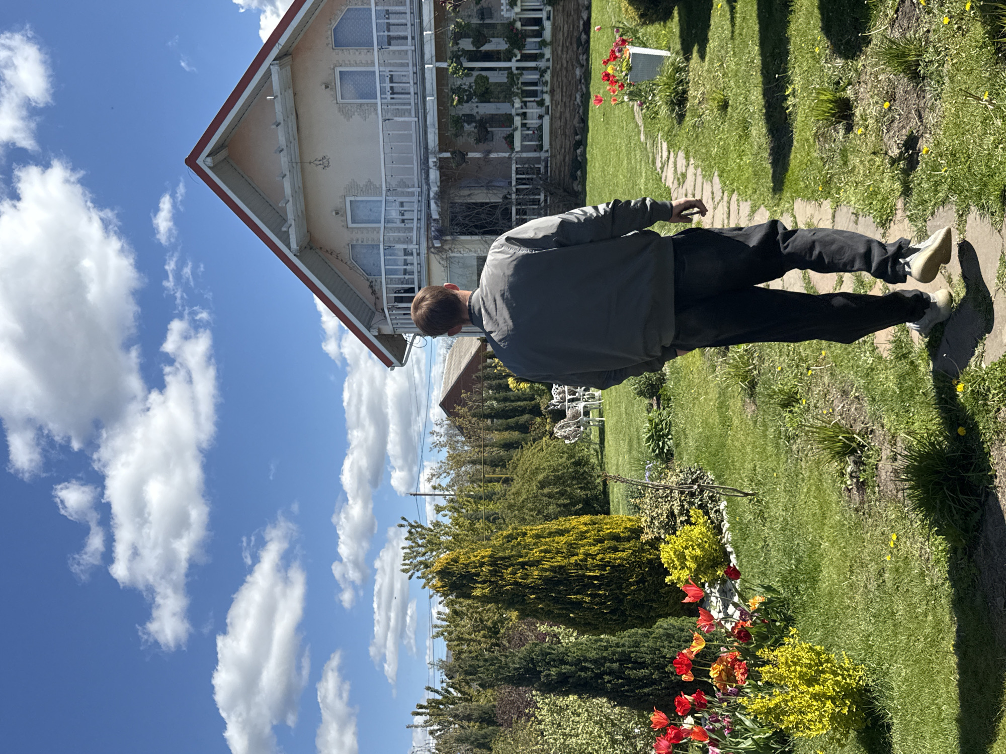 This vertical image depicts a sunny garden scene in what appears to be a suburban or rural setting, likely in Pervorechenskoye, Russia.
The primary subject is a person, seen from behind, standing on a winding stone path. The person is wearing a dark grey jacket, dark trousers, and light-colored shoes. They are looking towards a two-story house with a light-colored facade, multiple windows, and extensive white balconies on both floors. The house has a distinctive red-trimmed gable roof.
The surrounding environment is a vibrant garden, featuring a neatly mown green lawn, various trees, and shrubs. Brightly colored flowers, predominantly red and yellow tulips, are visible in flower beds lining the stone path. In the background, beyond the house, more trees and greenery extend, and some white garden furniture can be glimpsed.
The activity taking place is minimal; the person appears to be observing the house or garden, possibly strolling through it.
Notable details include the bright, clear blue sky dotted with scattered white cumulus clouds, indicating fair weather. The strong sunlight casts distinct shadows, suggesting it is daytime, likely mid-morning or afternoon. The overall impression is one of tranquility and natural beauty. There is no visible text in the image.