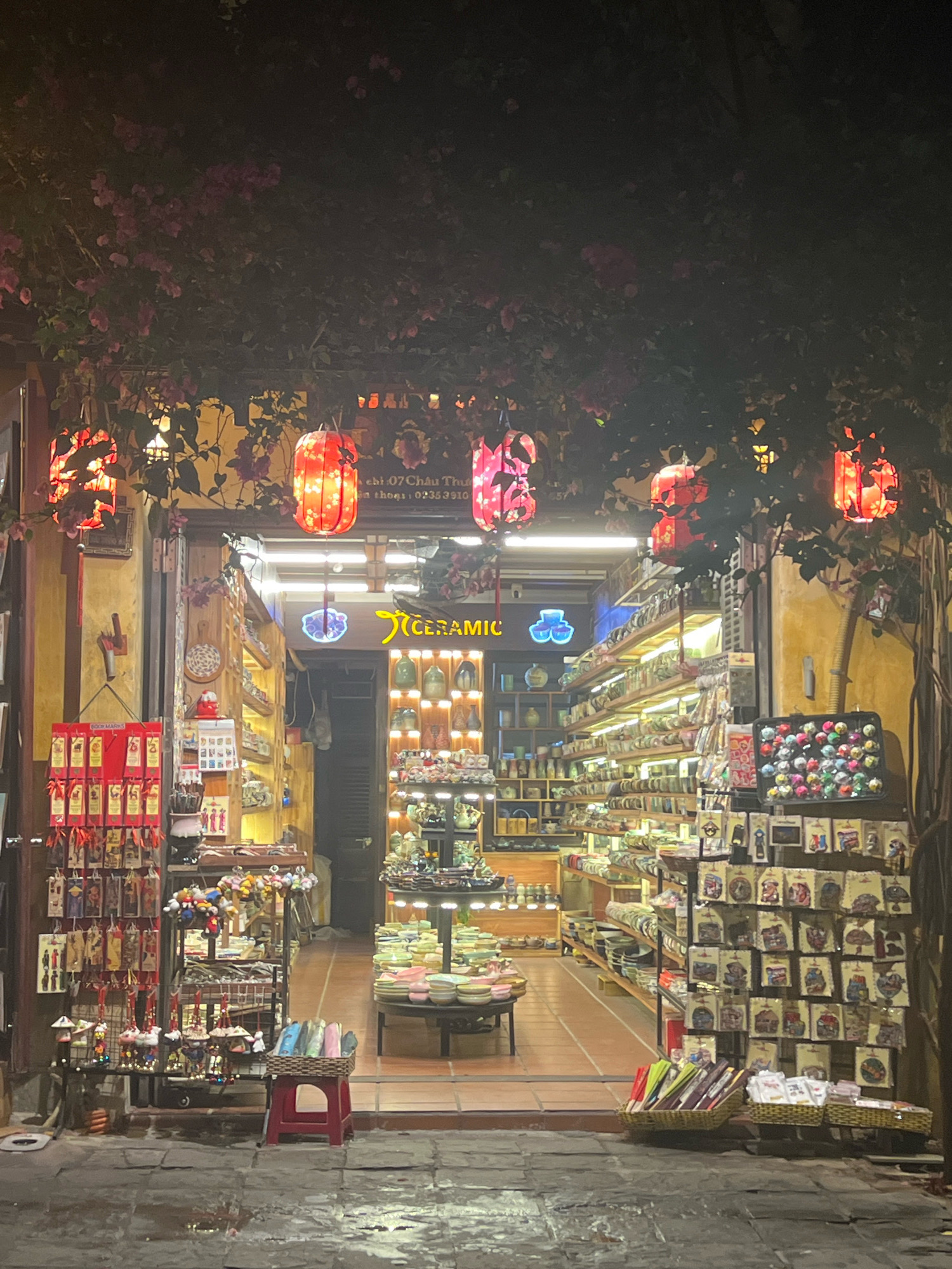 The image depicts the entrance of a shop at night in Hoi An, Vietnam, likely on a street lined with similar establishments. The shop, named "CERAMIC" as indicated by a neon sign, specializes in handcrafted items. Red lanterns hang overhead, casting a warm glow on the street and illuminating the facade of the building. The shop's interior is visible, displaying a wide array of ceramic goods, including vases, bowls, and decorative items, arranged on shelves. In front of the shop, various trinkets and souvenirs are displayed on tables and racks, such as colorful tassels, bookmarks, and small figurines. The ground in front of the shop appears wet, suggesting recent rain. The overall atmosphere is inviting and colorful, typical of the vibrant night markets and shopping streets in Hoi An. There are no people visible in the image.