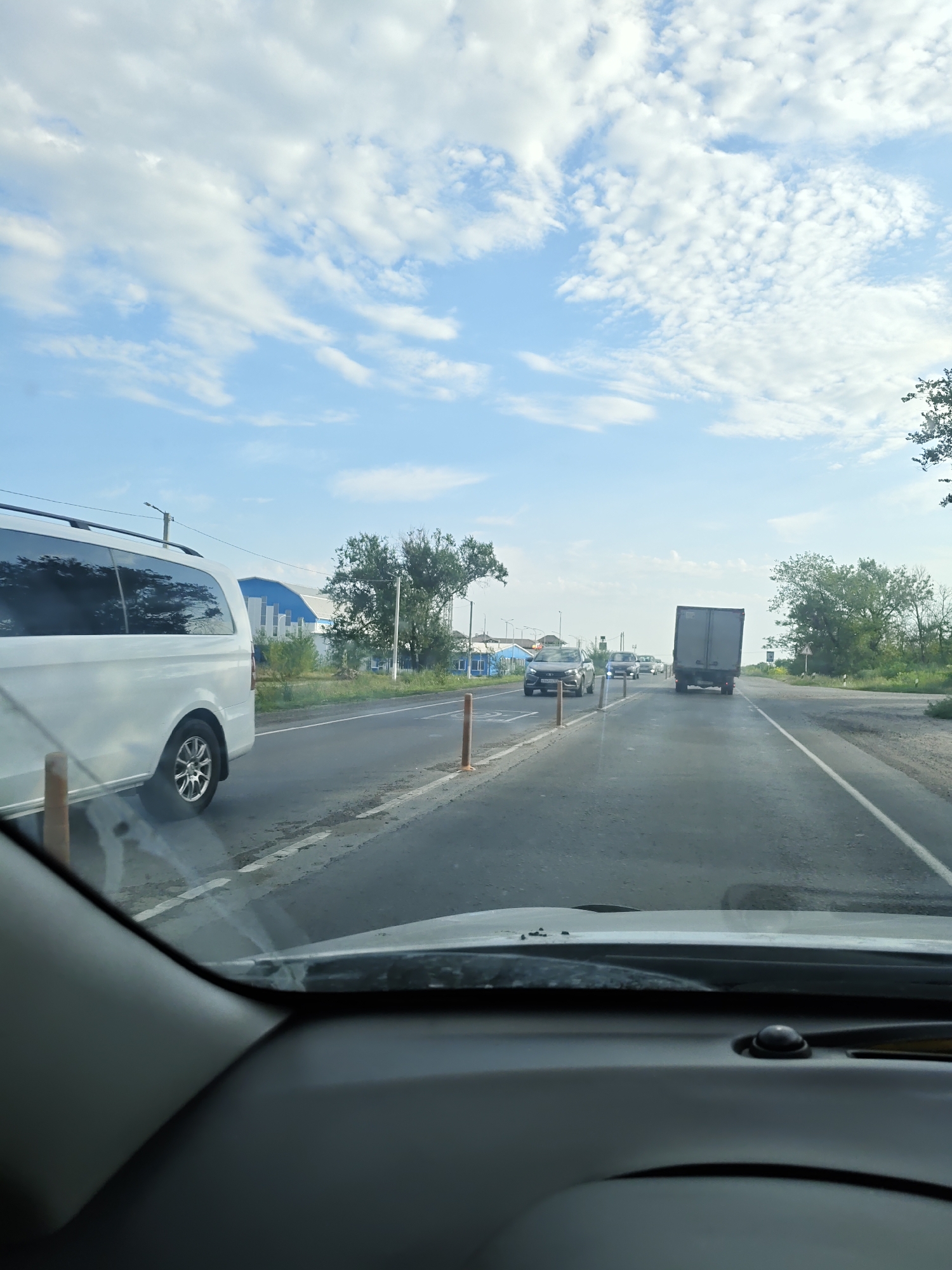 The image shows a view from inside a car, looking out at a two-lane road on a cloudy day. In the foreground, the car's dashboard is visible. The road is lined with orange traffic cones. On the left side of the road, a white van is driving. Further down the road, there are several other vehicles, including a truck and some cars. On the right side of the road, there are trees and some buildings. The sky is blue with scattered white clouds. The scene appears to be a rural or suburban area in Russia. The time of day appears to be daytime. There are no people clearly visible in the image. The overall mood is neutral, depicting a typical travel scene. There is no visible text in the image.