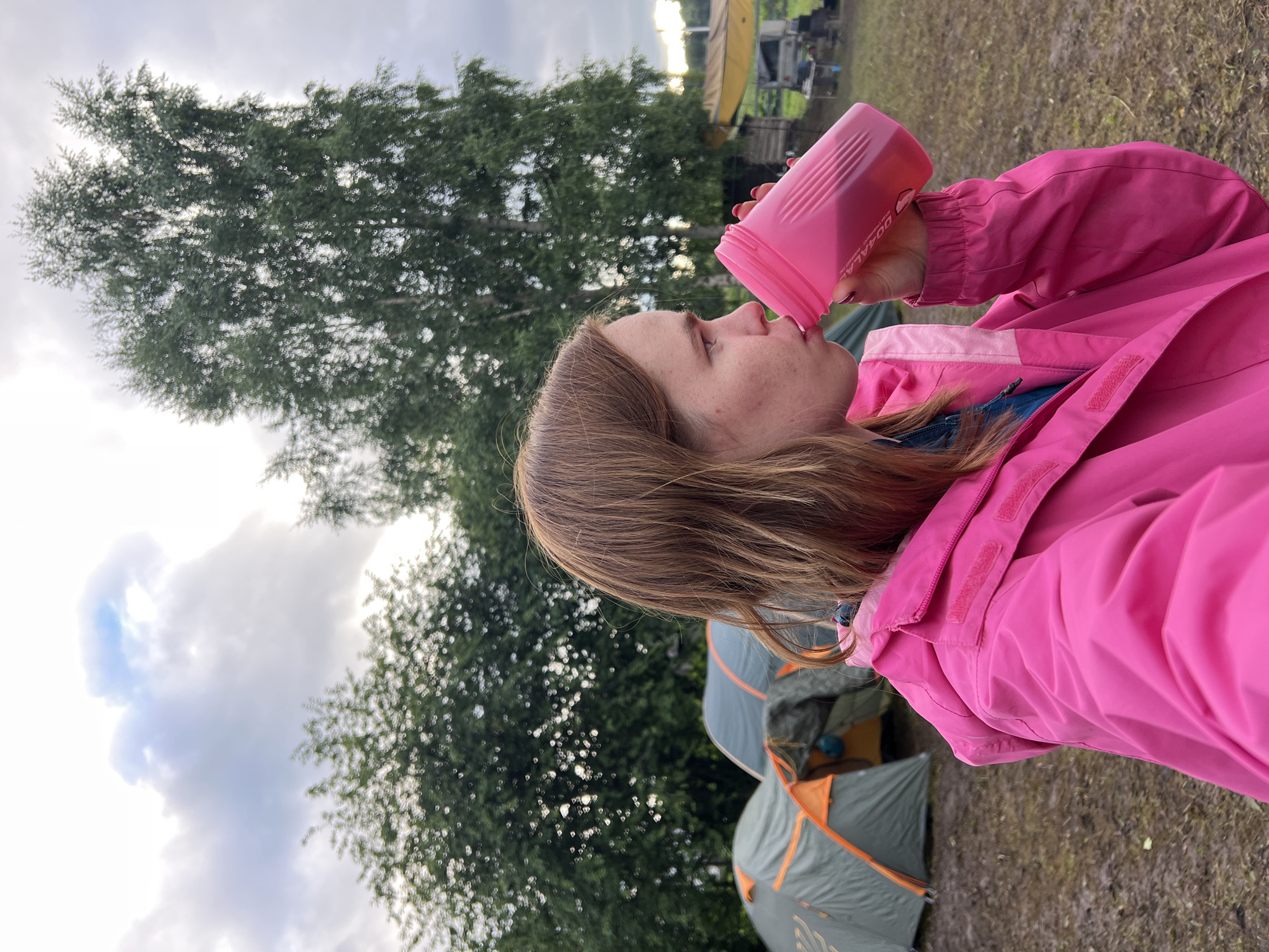 A young woman with brown hair, wearing a bright pink jacket, is taking a drink from a pink water bottle. She is seen in profile, with the water bottle raised to her mouth. The background of the image shows a campsite with several tents, indicating an outdoor setting. Tall green trees are visible behind the woman and tents, and the sky above is overcast with clouds, suggesting potentially cooler weather. The overall scene appears to be at a campsite, possibly during the daytime given the ambient light. The woman's expression is neutral as she drinks. Text is visible on the water bottle: "OOALA".