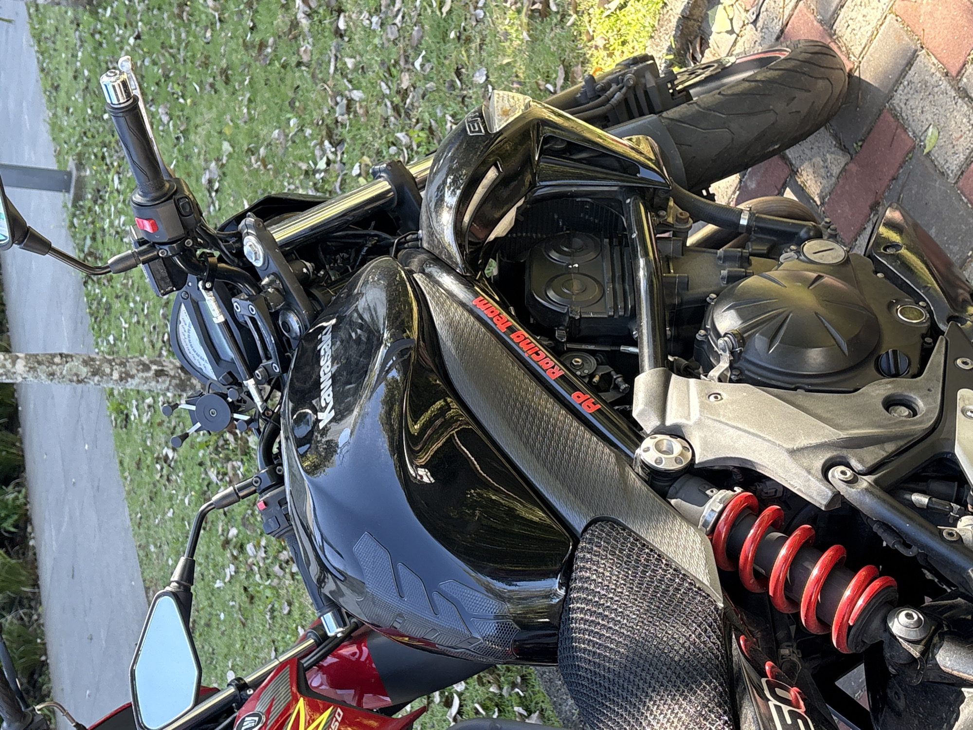 This is a medium shot, taken from a slightly low angle, of a black Kawasaki motorcycle parked on a brick pathway in what appears to be a park or garden area. The motorcycle is the primary subject, and its details are clearly visible. The front of the motorcycle is on the right side of the frame, with the rear on the left.
The motorcycle is predominantly black, with a glossy fuel tank that reflects the surrounding environment, showing trees and sky. There is a textured black decal on the top of the fuel tank. A prominent red coil spring is visible on the rear suspension, adding a splash of color to the otherwise dark machine. The seat appears to be a textured black material. On the side of the fuel tank, there is a red decal with white lettering that reads "AP RACING TEAM". Part of a red motorcycle is visible on the far left of the frame, suggesting another vehicle is parked nearby.
The background consists of a grassy area with scattered fallen leaves. Behind the grass, there are trees and foliage, suggesting an outdoor setting. The ground beneath the motorcycle is paved with red and brown bricks, laid in a patterned walkway. The lighting indicates it is daytime, with sunlight casting shadows on the ground and highlighting the motorcycle's surfaces. No people are visible in the image.
Based on the visual cues, such as the vegetation and pathway style, and the specific context of the motorcycle branding, it is plausible this image was taken in a city park within São Paulo, Brazil. For instance, the Ibirapuera Park often features similar brick pathways and diverse greenery, and the presence of a custom-styled motorcycle is not uncommon in urban areas of São Paulo where motorcycle culture is prevalent. The "AP Racing Team" sticker might indicate a connection to local racing or custom bike communities that exist in and around São Paulo.