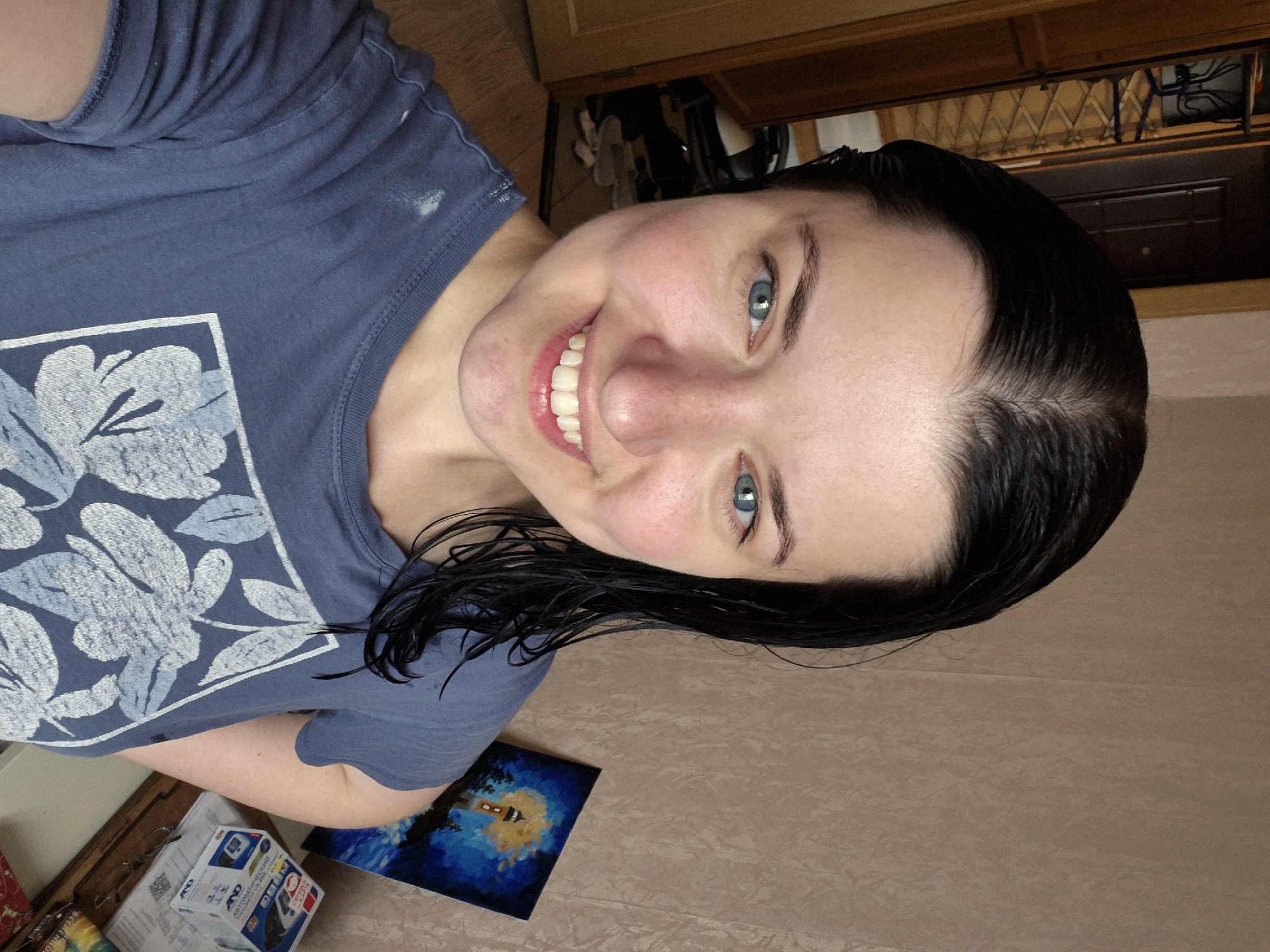 A young woman with black hair and blue eyes, wearing a blue t-shirt with a floral print, smiles at the camera. Her hair appears wet and styled to one side. She is standing in what appears to be an indoor setting with a door and part of a closet visible in the background. A small painting of a lighthouse is on display on a surface to her right. The overall lighting suggests an interior space. This image does not contain enough information to determine a specific location in Saint Petersburg, Russia.