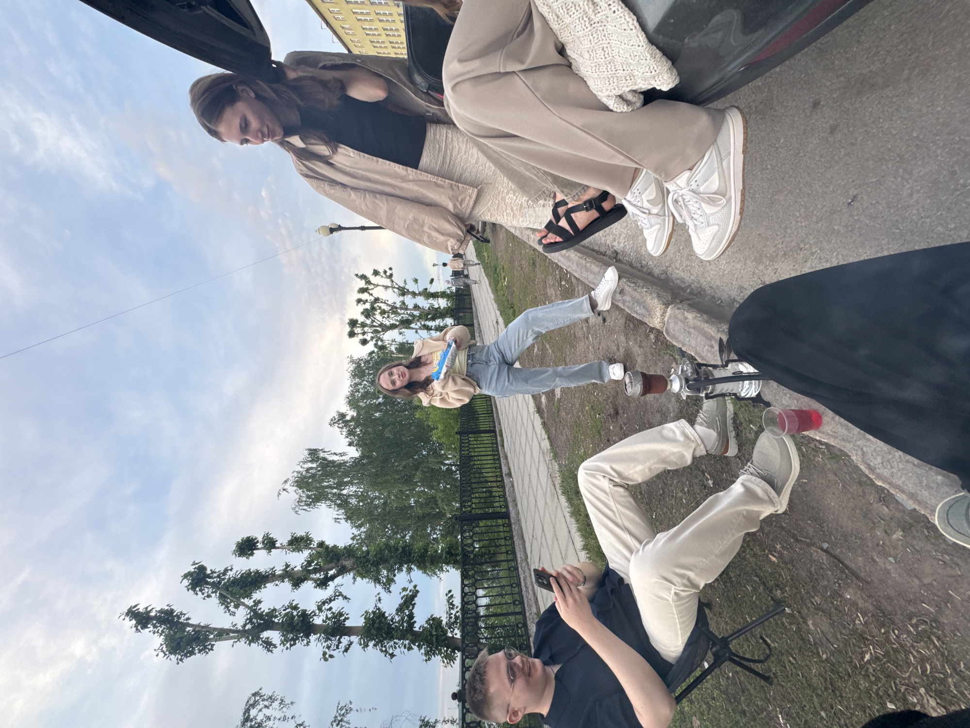 The image shows a group of young people gathered outdoors in an urban setting. Two women are sitting in the open trunk of a dark-colored car, parked along the curb. One woman is wearing a black top with a beige jacket and patterned pants, while the other is wearing beige pants and white sneakers. Another young woman is sitting on the curb, wearing a beige hoodie and jeans. Two young men are also present. One is seated on a camping chair, looking at his phone. He is wearing a dark t-shirt and light pants. Another man is sitting nearby. There is a hookah present. The location is likely in Perm, Russia, due to the architectural style of the buildings in the background. The group is enjoying the outdoor gathering, suggesting a casual and social atmosphere. The weather appears clear and sunny.