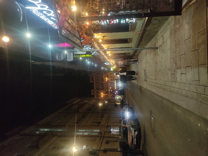 The image depicts a street scene at night in Moscow, Russia. The street is lined with buildings, some of which have illuminated signs and decorated facades. Pedestrians are visible walking along the sidewalk. A car with its headlights on is traveling on the road. Streetlights provide additional illumination. The pavement appears to be made of paving stones.
