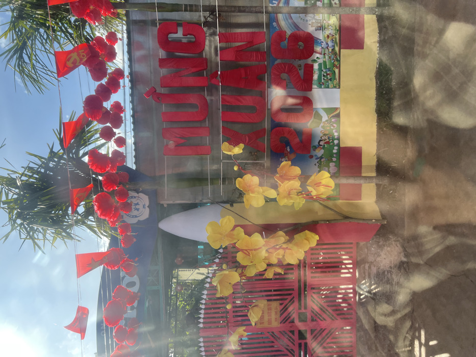 The image depicts an outdoor decorated facade, likely an entrance, in the city of Xuân Long, Vietnam, under bright daylight conditions. The foreground exhibits significant reflections from a glass surface through which the photograph was captured. Overhead, a string of red triangular pennants, some featuring a yellow star (Vietnamese flag design) and others displaying a yellow hammer and sickle symbol, are strung between two prominent palm trees against a clear blue sky. Numerous red spherical or flower-like decorations are suspended alongside the pennants. Below these, a gray corrugated metal roof is visible.
To the left, a red metal gate with vertical bars is partially obscured by artificial yellow apricot blossoms ("mai" flowers) arranged on a tall, conical structure colored white and yellow. A small white sign with red text, partially legible as "Tự Yêu Cầu..." and "Khi Vào Sân," is affixed to the gate. Dominating the center-right is large red Vietnamese text spelling "MỪNG XUÂN 2026" (Happy Spring/New Year 2026). Beneath the numerical "2026," colorful murals or posters illustrate groups of children playing in various outdoor settings, featuring elements like a rainbow and playground equipment. The wall supporting the signage displays sections of red and yellow paint. No individuals are present in the visible scene.