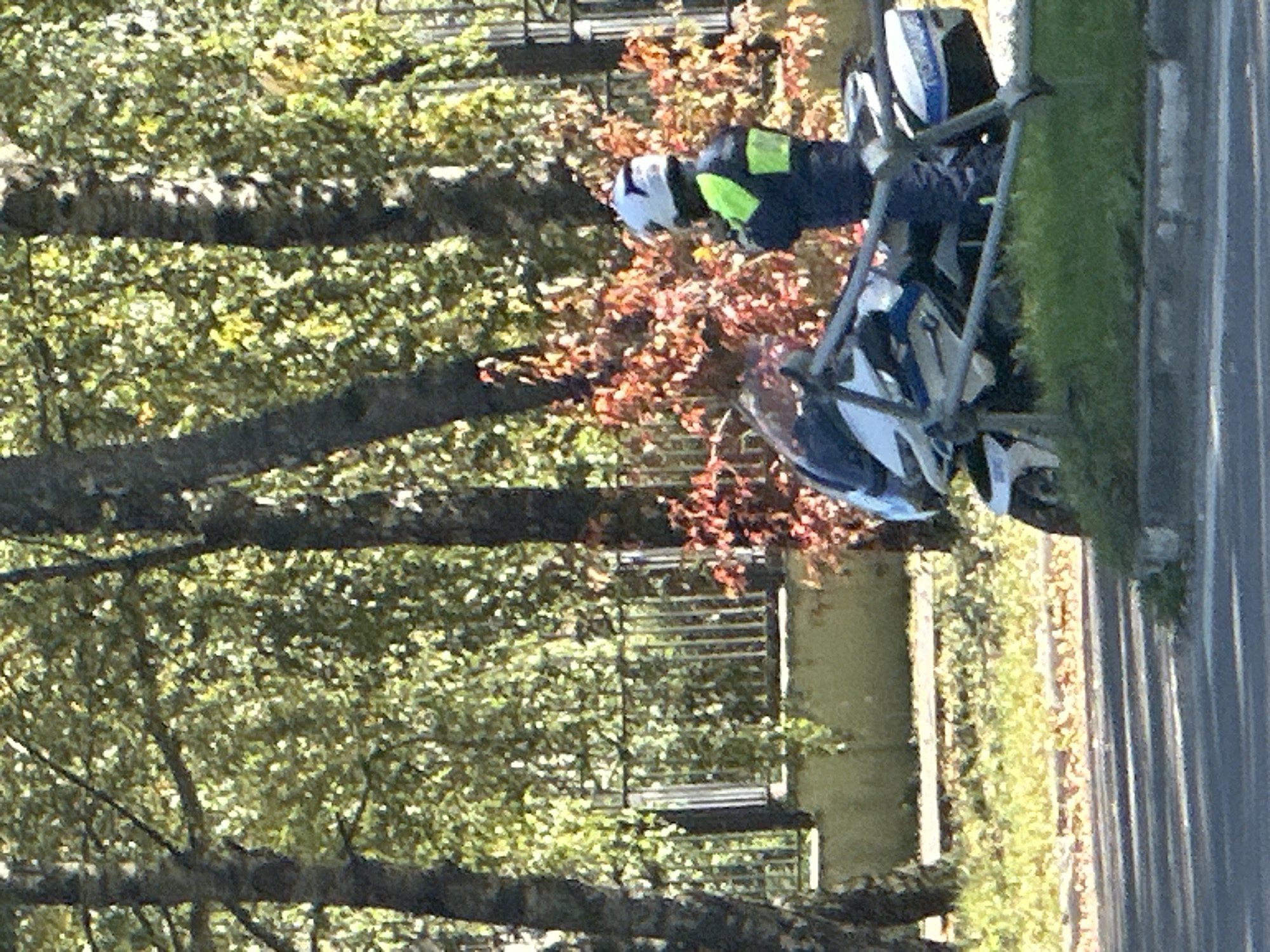A person wearing a white helmet and a high-visibility jacket over a dark uniform stands next to a white police motorcycle. The motorcycle has blue and white markings and is parked near a metal barrier. The scene is outdoors, with trees and foliage in the background, suggesting a park or roadside setting in Podolsk, Russia. Sunlight illuminates the area, casting shadows.