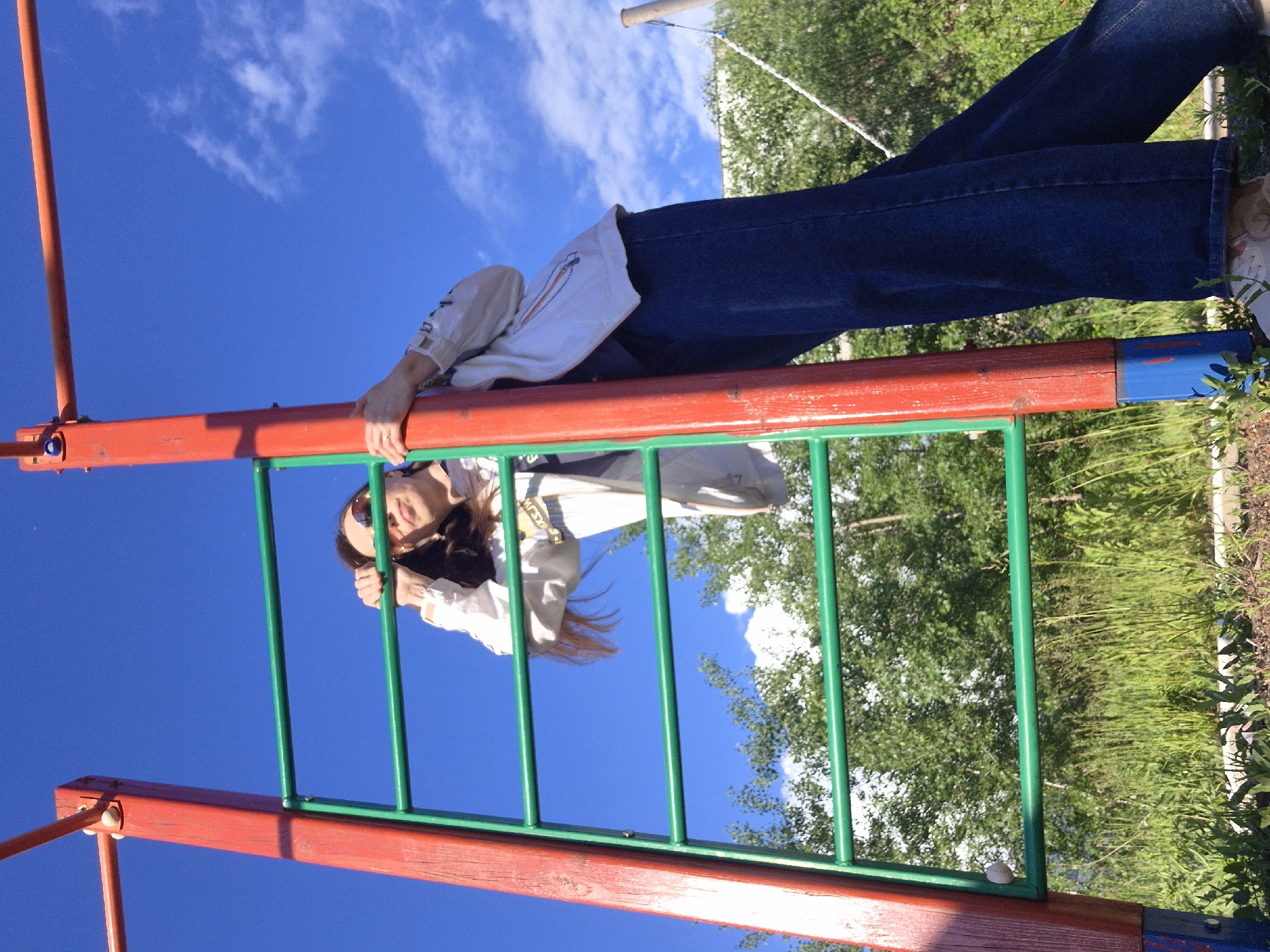 A young woman is depicted hanging upside down from a red horizontal bar of a playground structure in an outdoor setting in Chaikovsky, Russia. The photograph is oriented 90 degrees clockwise, causing the clear blue sky with sparse white clouds to appear on the left, and green foliage, trees, and grass on the right.
The woman is wearing a light-colored (possibly white or off-white) long-sleeved jacket or top, blue jeans, and light-colored sneakers. She has dark hair, which is flowing downwards due to gravity, and is wearing sunglasses. Her head is positioned towards the bottom-left of the frame, and her feet are towards the top-right, with her body suspended from the bar by her arms and torso. She is looking towards the viewer with a neutral or slight smiling expression.
The playground equipment features prominent red wooden or metal bars and green metal bars, showing some signs of wear and chipping paint. The bright lighting indicates a sunny day. In the background, beyond the playground structure, there is an abundance of green foliage and trees, characteristic of a park or natural outdoor area. No legible text is visible on the woman's clothing or the playground structure.