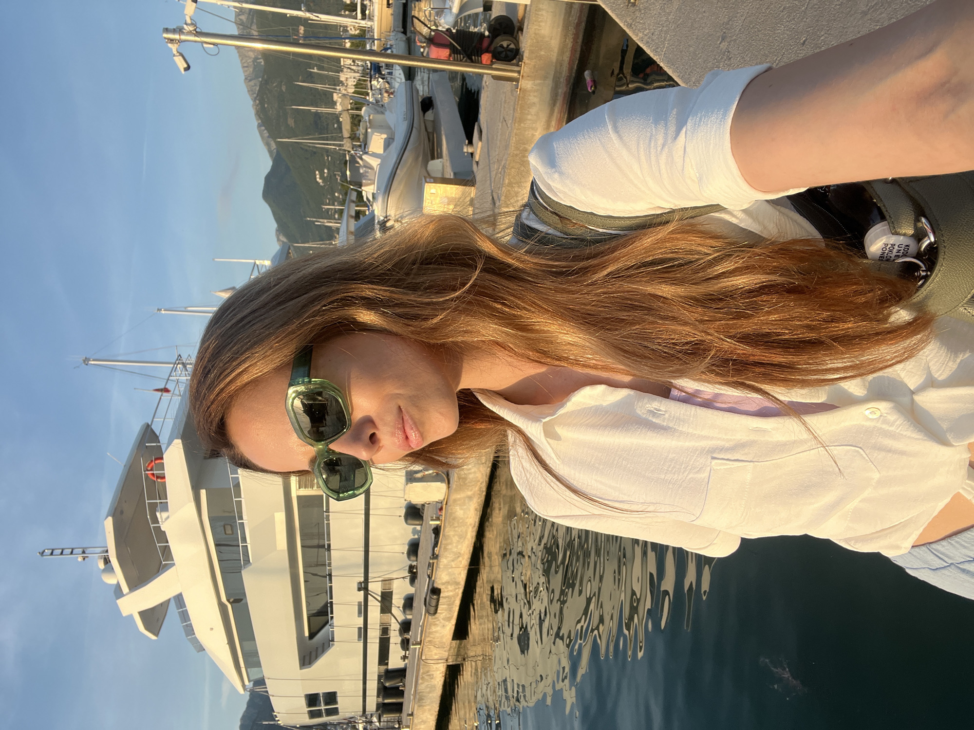A sunlit portrait captures a woman standing at a marina in Bar, Montenegro, during the golden hour of late afternoon or early evening. The warm, directional sunlight illuminates her and the surrounding environment, suggesting clear, pleasant weather.
The woman, positioned on the right side of the frame, is seen from the chest up. She has long, flowing reddish-brown hair, brightly lit by the sun. She wears stylish green-framed sunglasses and a white, loose-fitting button-up shirt over a lighter-colored top, possibly light blue or purple. Her expression is relaxed, with her lips slightly parted. A dark green bag or backpack rests on her shoulder, from which a white tag or keychain is visible, displaying partial text that reads "KOD KUPUJE PONE..." or "KODKUPI...".
The background reveals a vibrant marina with numerous boats docked along a concrete pier. These include various sailboats with their masts soaring into the blue sky, and a prominent large, white motor yacht with multiple decks and windows. The calm water of the marina reflects the sky and boats, showing subtle ripples. In the distance, majestic mountains rise under a clear blue sky, forming a scenic backdrop to the coastal setting. The scene evokes a tranquil and picturesque moment by the water.