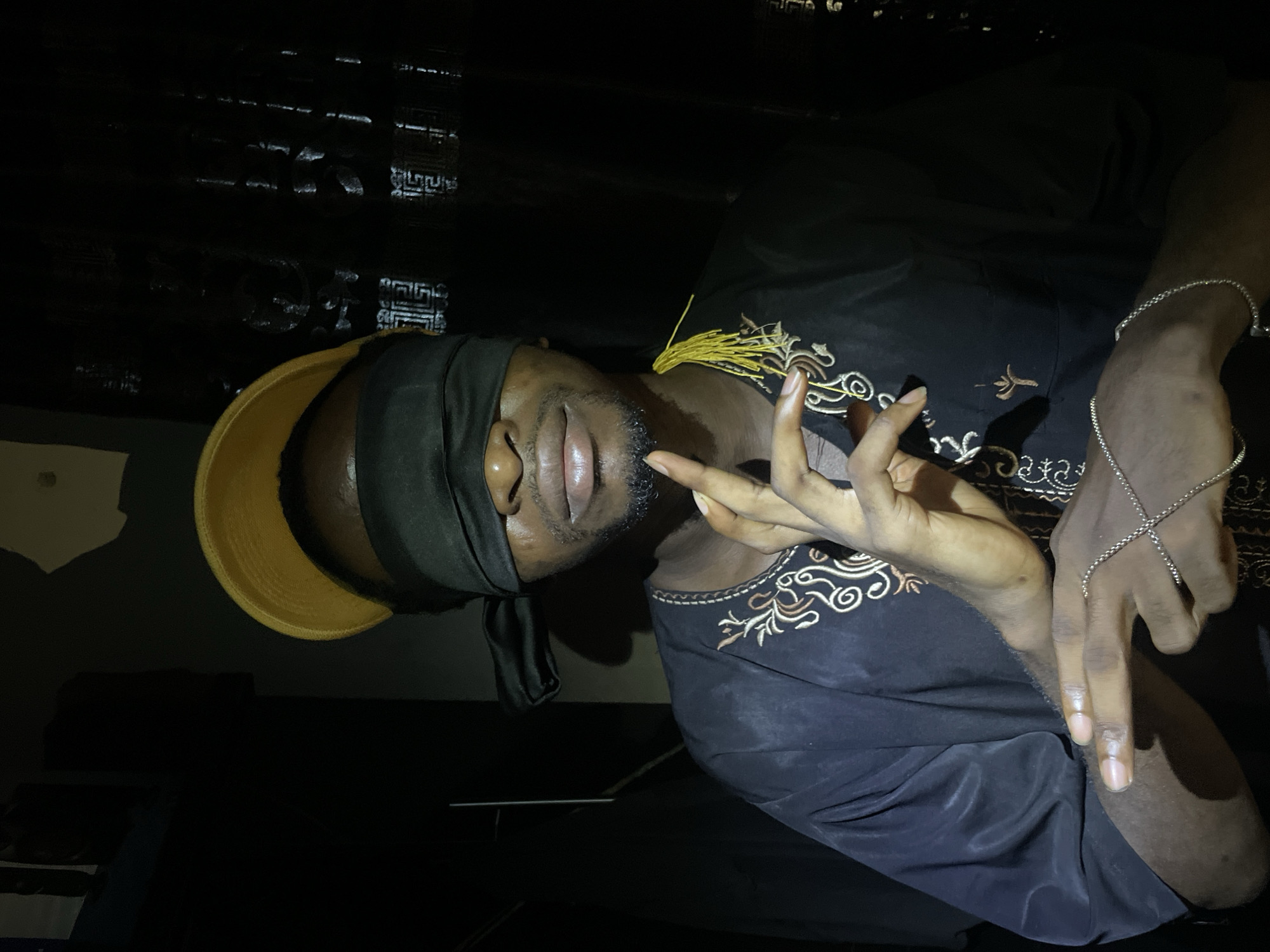 A man is shown from the chest up, with a black cloth covering his eyes. He is wearing a black garment with gold embroidery. He is wearing a gold and black hat and two silver bracelets. He is making a gesture with his hands. The lighting is dim. The location is likely indoors but is indiscernible. The city is Lokoja, Nigeria, but there are no details in the photo to confirm the location.