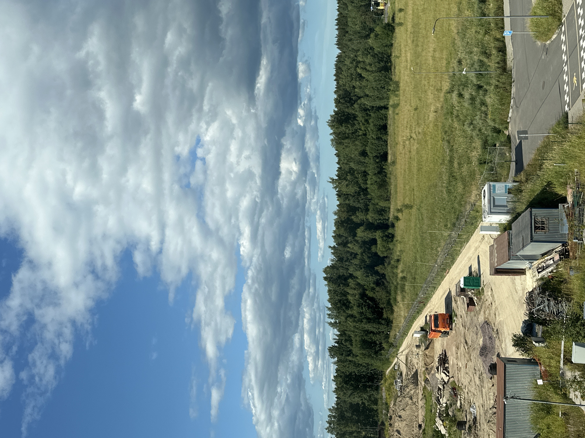 The image depicts a construction site in Vsevolozhsk, Russia, under a cloudy sky. In the foreground, there are several corrugated metal structures and piles of debris. A dirt road leads into the site, where an orange truck is parked. Further in the background, a dense forest borders a grassy field. The scene appears to be in a semi-rural or industrial area on a clear day with scattered clouds.