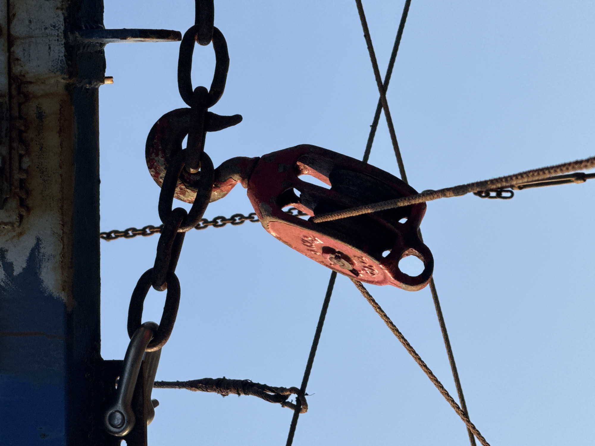 The image depicts a rusty, weathered pulley system against a clear, pale blue sky. The system is connected to a thick, dark metal support structure with visible paint chipping and rust. A heavy chain is connected to the support and extends down to a shackle. The pulley itself is painted red, though the paint is faded and worn. A rope is threaded through the pulley, extending to the right. Additional ropes and chains are also present. The location in the city of Emona, Bulgaria, is not discernible from the image.