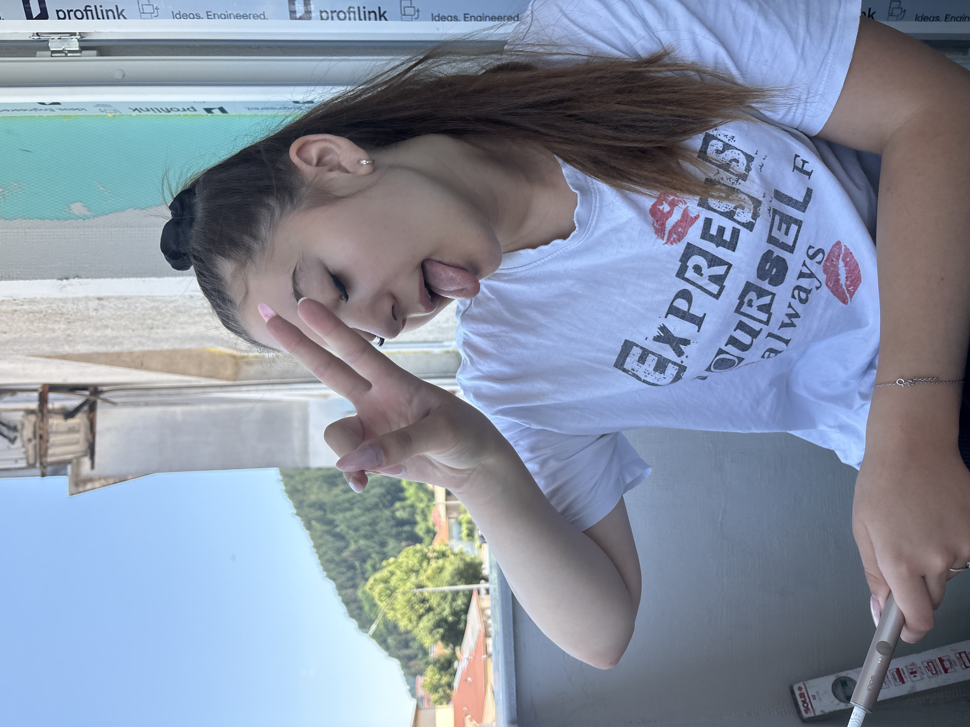 A young woman with long, brown hair is pictured in a close-up shot, sticking her tongue out and making a peace sign with her right hand. She is wearing a white t-shirt with the words "EXPRESS YOURSELF" and kiss marks printed on it. A black scrunchie is visible in her hair. The background shows a bright blue sky with some greenery and buildings in the distance, suggesting an outdoor setting. The image appears to be taken during the daytime, with clear weather. Text visible on a surface in the lower part of the image includes "profilink" and "Ideas. Engineered." Additionally, the word "iQOS" is visible on a device she is holding. The overall mood is playful and casual.