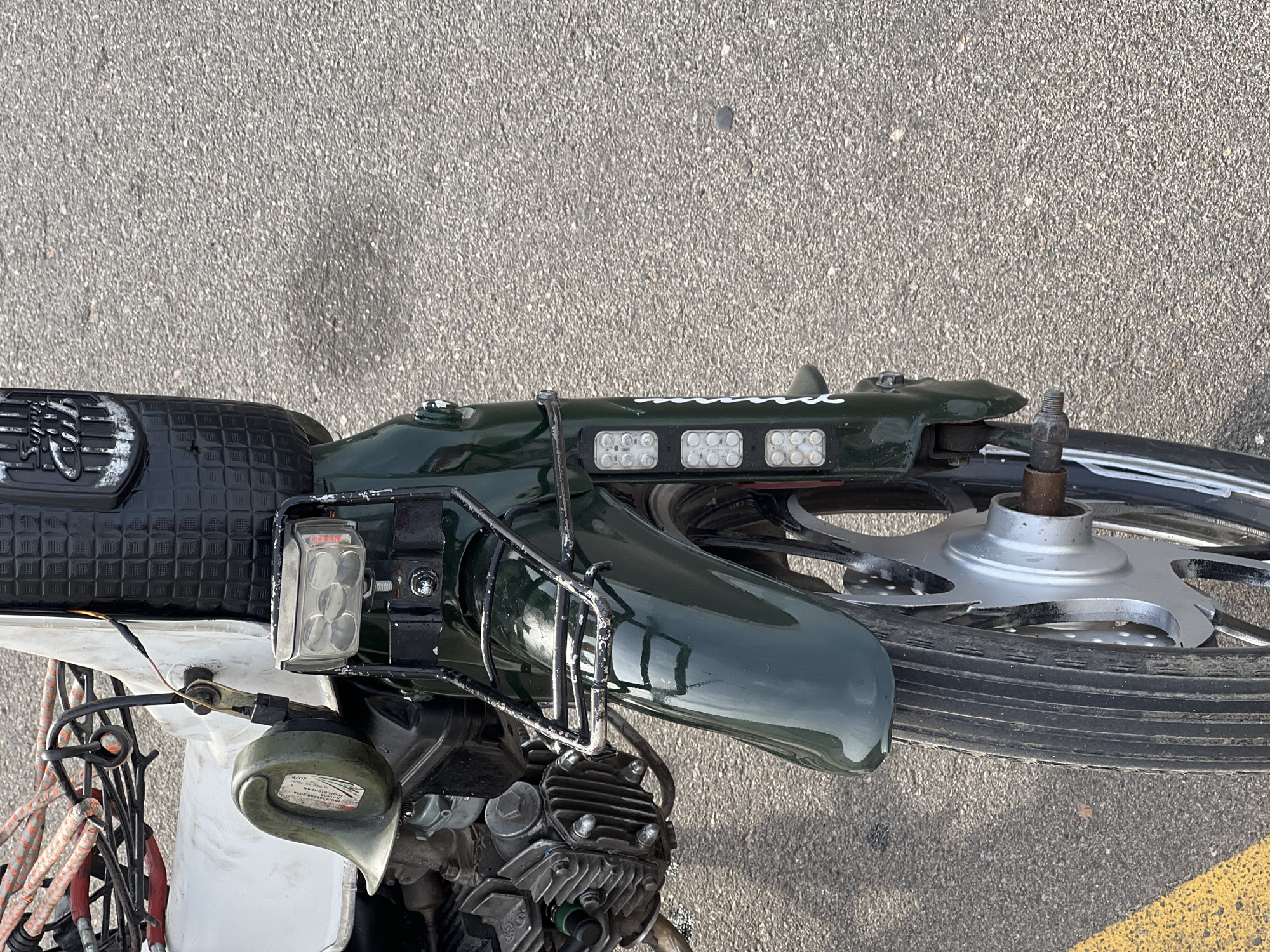 An image taken in Jalingo, Nigeria shows the front of a green scooter parked on an asphalt road with a yellow line on the left side. The scooter features a black horn on the left side, a rectangular headlight with multiple bulbs, and three vertical LED lights on the front fender. Part of the engine and a white panel are also visible. The background is a textured asphalt surface.