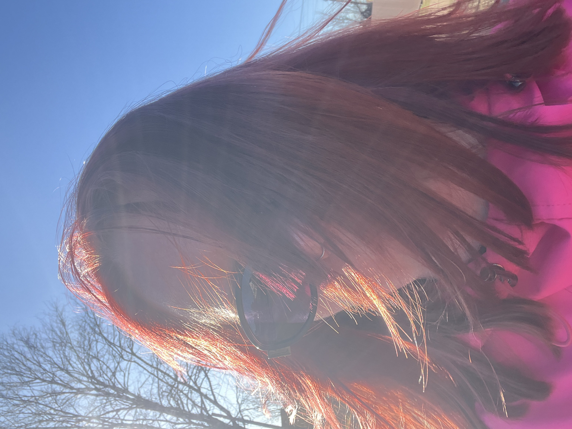 The image shows a person with long, reddish-brown hair and wearing sunglasses and a pink jacket. The photo appears to have been taken outdoors on a sunny day, as evidenced by the blue sky and bright sunlight. Bare tree branches are visible in the background. There is no information in the image to confirm it was taken in Chaikovsky, Russia.