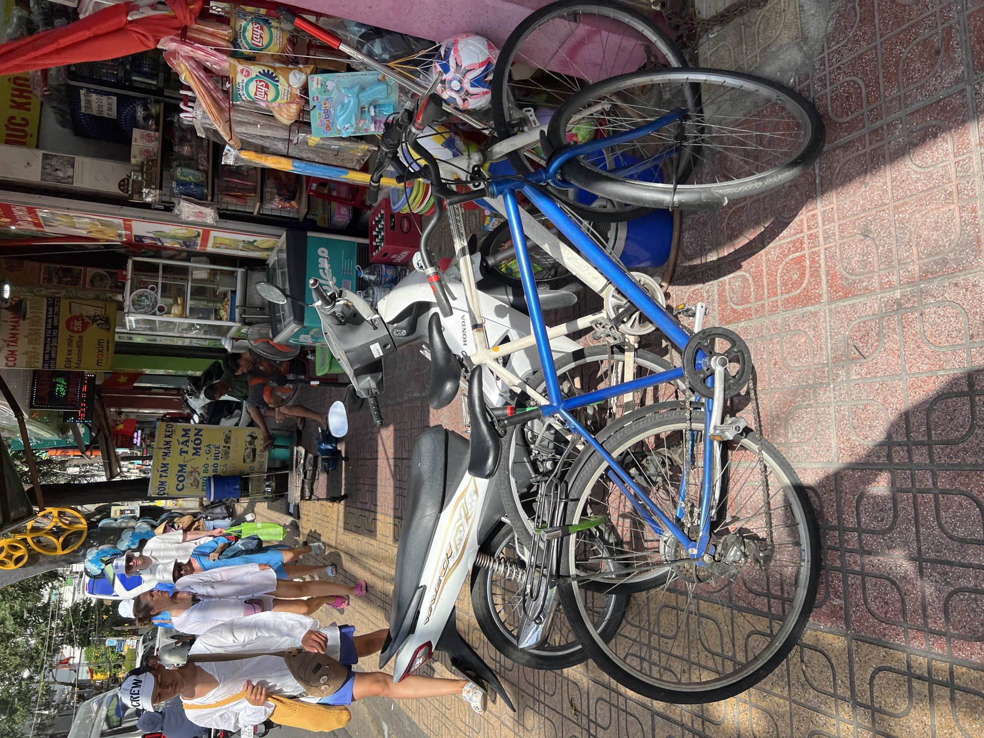 In Nha Trang, Vietnam, a sun-drenched street scene unfolds, featuring a bustling sidewalk lined with shops and parked vehicles. A white scooter is prominently parked, its polished chrome reflecting the sunlight. Next to it, a blue bicycle with a silver frame leans against a wall, its spokes catching the light. In the foreground, the intricate patterns of the tiled sidewalk are visible, casting long shadows from the overhead sun.
Further down the street, a group of people are walking, dressed in casual attire. One individual, wearing a white cap and a light-colored shirt, carries a beige woven bag slung over their shoulder. They are accompanied by a woman in a white shirt and a blue dress, and another person in a white shirt and a white hat. The background reveals the vibrant commercial district, with shopfronts displaying a variety of goods, including colorful snacks and beverages. A sign in Vietnamese is visible on one of the shop fronts, advertising food. The scene captures a typical day in this lively Vietnamese city, with a mix of pedestrians, vehicles, and commercial activity.
