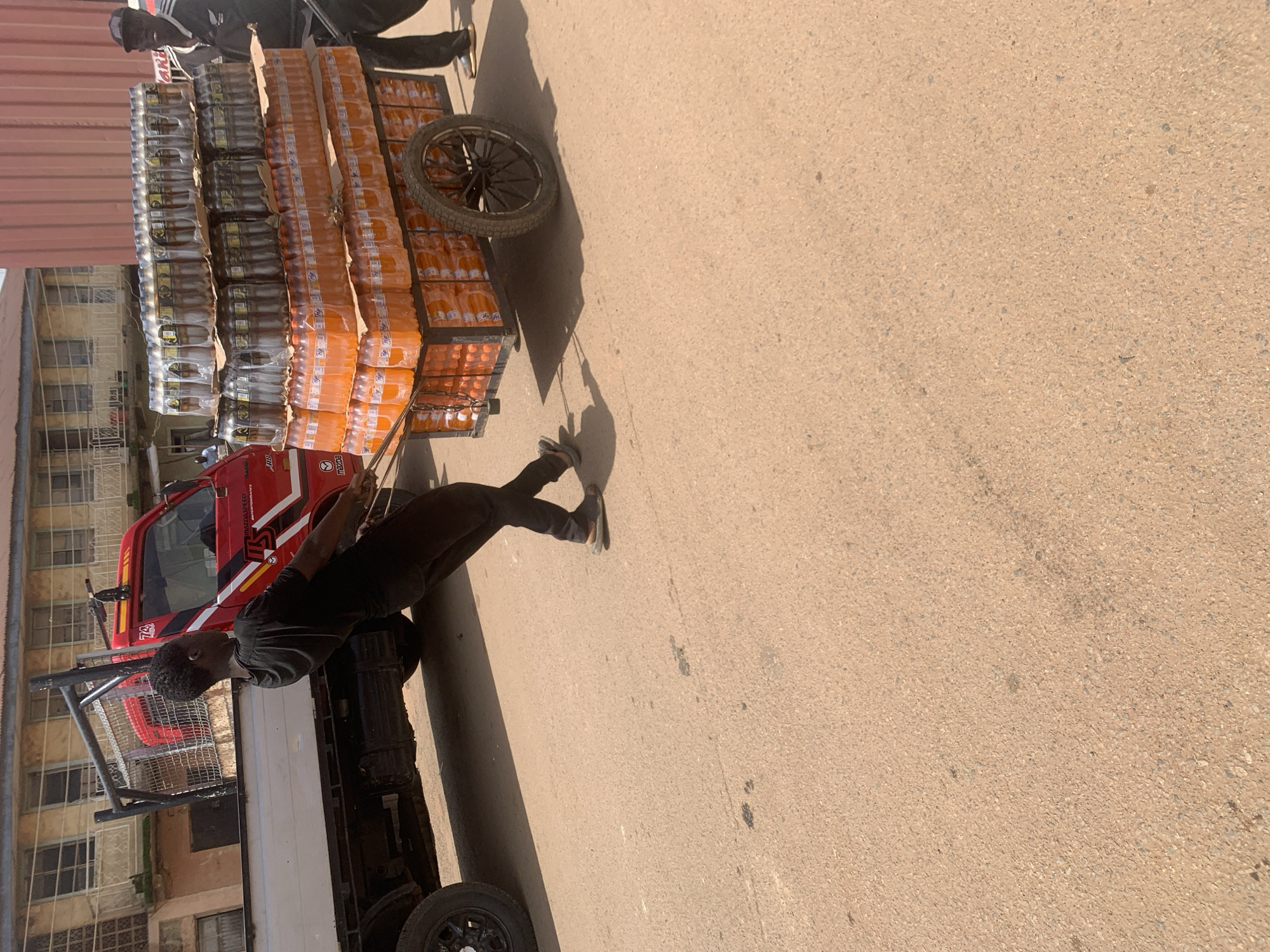 This media file captures an outdoor scene in Jos, Nigeria, likely during the daytime given the strong shadows. The primary subjects are a man and a cart loaded with beverages. The man, dressed in a black t-shirt and dark pants, appears to be interacting with the cart or preparing for its movement. His posture suggests he might be pushing, pulling, or adjusting the load.
The cart is a prominent object, heavily laden with stacked rows of bottled beverages. The bottles are predominantly orange, suggesting they contain a soft drink like Fanta, and are shrink-wrapped in clear plastic for stability. The cart itself has a utilitarian design, with a metal frame and a visible wheel, indicating it's likely manually operated. To the side of the cart, a red vehicle, possibly a small truck or utility vehicle, is partially visible. Its presence suggests a transport or delivery operation.
The scene unfolds on a paved surface, likely a street or market area, with a building visible in the background. The overall impression is one of a typical street-side commercial activity. The lighting indicates it is a sunny day, casting sharp shadows. The general atmosphere appears to be one of everyday commerce and labor, with no overt display of strong emotions. No visible text is clearly legible in the image.