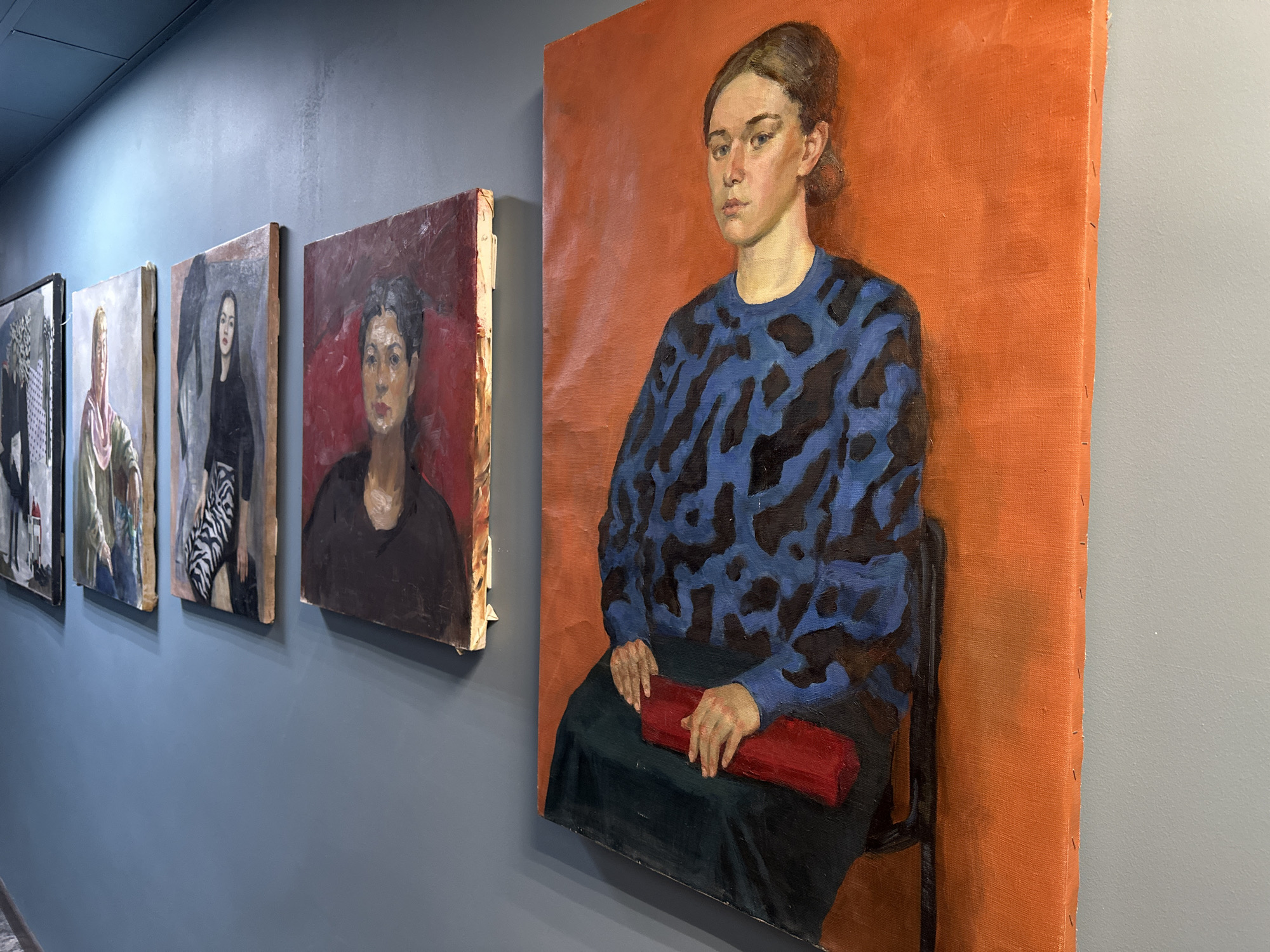 The image shows a series of oil paintings hanging on a gray wall in a room. The paintings appear to be portraits of various individuals. The painting on the far right is a portrait of a woman in a blue and black patterned sweater, seated on a chair, against an orange background. She is holding a red rectangular object in her lap. To the left are several other portraits, including one in dark colors, and others with lighter palettes. The specific location in Saint Petersburg, Russia cannot be determined from the visual information provided.