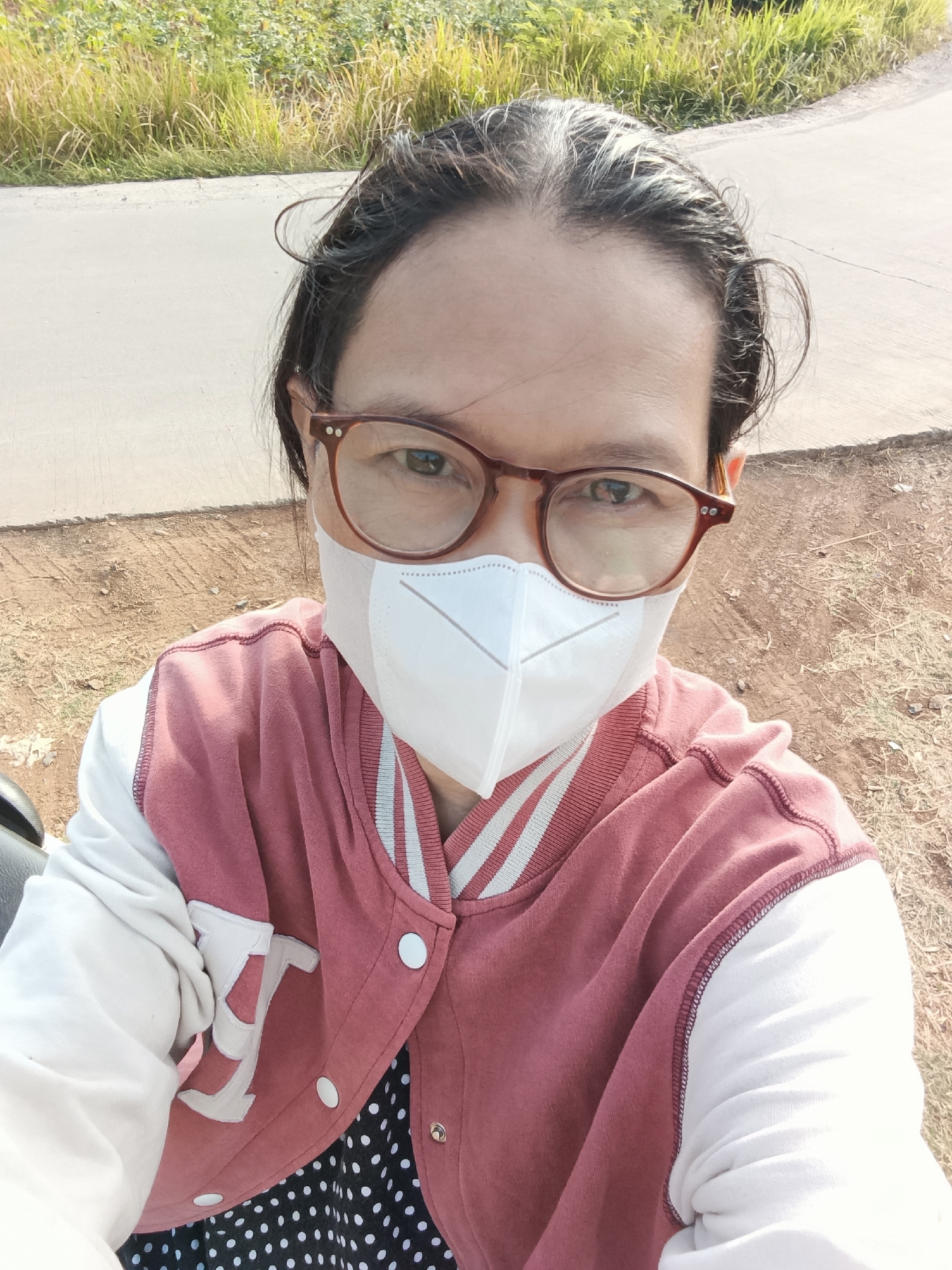 The image is a selfie taken outdoors, likely in Cluwak, Indonesia, given the context provided. The subject, a person wearing glasses and a white face mask, is the central focus. They are wearing a pink and white varsity jacket over a black and white polka dot shirt. The background shows a paved road with some dry vegetation and a hint of greenery, suggesting a rural or semi-rural setting. The lighting indicates it is daytime, with the sun casting shadows. The individual is looking directly at the camera, and their hair is dark and tied back. The overall impression is a casual, personal photograph.