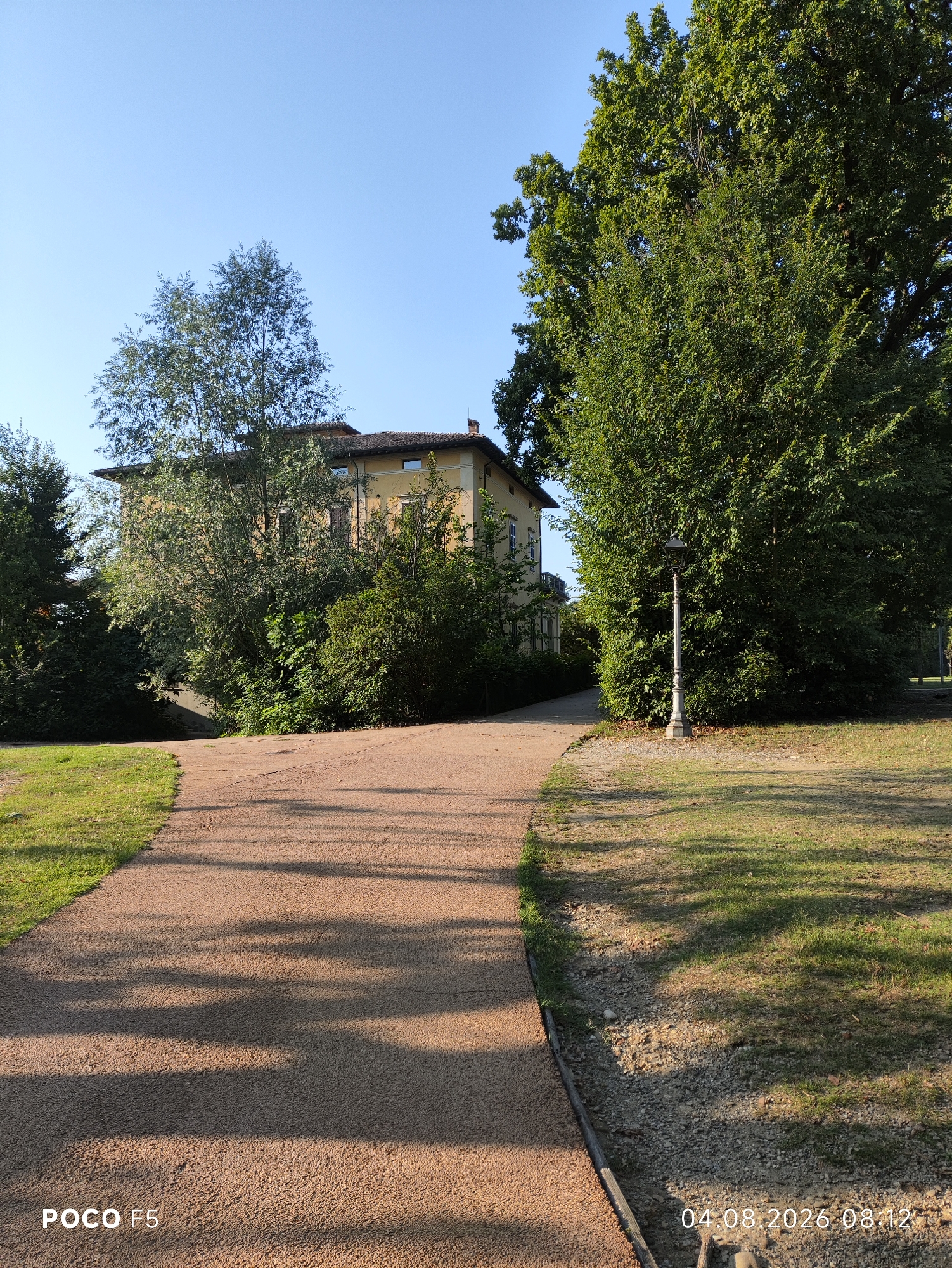 The image depicts a park setting with a building and a path. A two-story, pale yellow building with a red-tiled roof is partially visible behind a dense arrangement of trees and shrubs. The building has multiple windows and a chimney. In front of the building, a wide, light brown gravel path curves to the right, leading into the distance. To the left of the path is a grassy area. On the right side of the path, a tall, black lamppost stands on a stone base amidst lush green foliage. The background is dominated by a clear blue sky and the large canopy of a mature tree. The lighting suggests it is daytime, with shadows cast by trees across the path and grass. The lower left corner of the image contains the text "POCO F5", and the lower right corner displays the date and time "04.08.2026 08:12". The scene appears to be a public park or garden. Based on available information, this scene is located within the grounds of the Parco Regionale dell'Orecchiella, in the municipality of Collecchio, Italy.