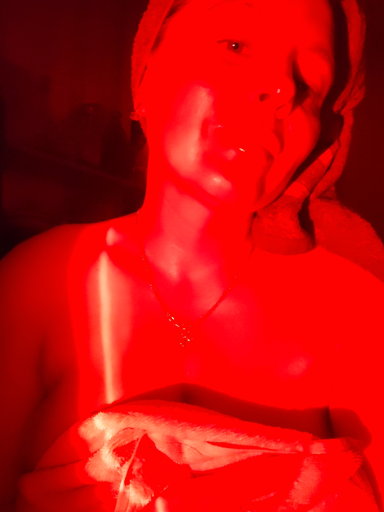 A person is seen from the chest up, illuminated by a red light. They are wearing a towel on their head and a towel draped around their shoulders, holding another towel in their hands. A necklace is visible on their neck. The background is dark and out of focus, with some faint shapes that could be furniture. No specific location in Izhevsk, Russia can be determined from the image.