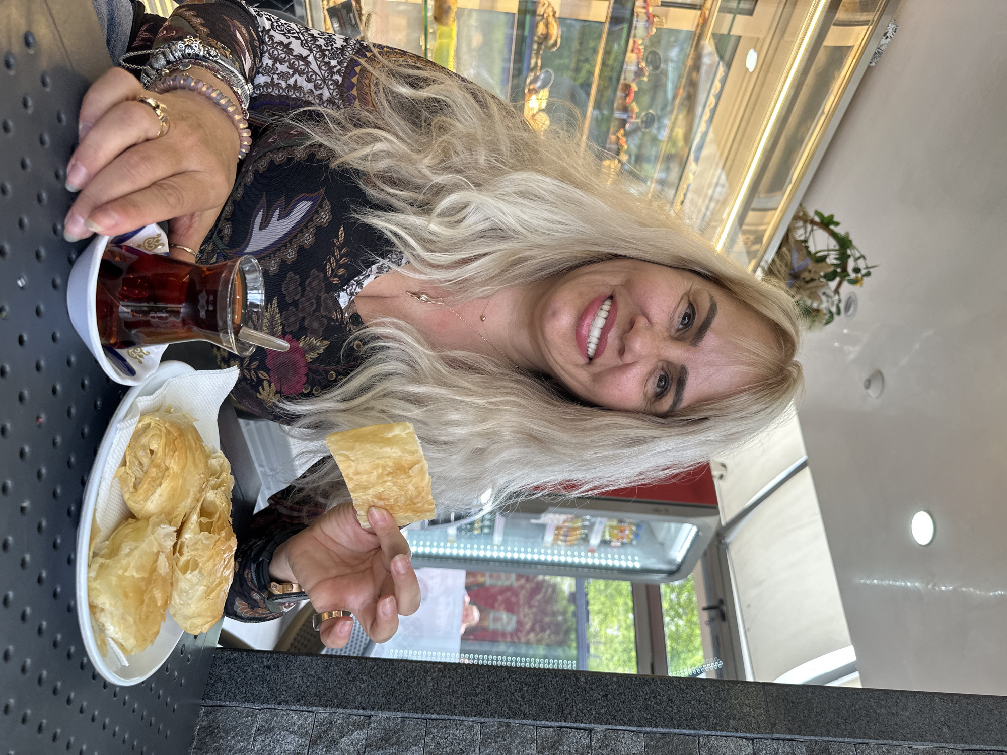 This image, taken in Sofia, Bulgaria, features a blonde woman smiling warmly at the camera while enjoying tea and pastries at what appears to be an outdoor cafe or bakery during the daytime.
The primary subject is a woman with long, wavy, light blonde hair and a bright smile. She is wearing a dark, long-sleeved top with an elaborate, lighter-colored pattern. Her left hand, adorned with multiple rings and bracelets, holds a clear, tulip-shaped glass of dark tea, resting in a white saucer with blue design accents. Her right hand holds a piece of golden-brown, flaky pastry. To her left, a white plate lined with a napkin holds several more identical pastries.
The scene suggests a relaxed setting at a food establishment. The woman is seated at a dark, textured or perforated table. In the background, a brightly lit display case with shelves holding various items is visible, indicating a commercial food setting. Beyond the display case, hints of an outdoor view with green foliage and natural light can be seen, possibly through a window or open-air section. The woman's expression conveys happiness and contentment. No discernible text is visible within the image.