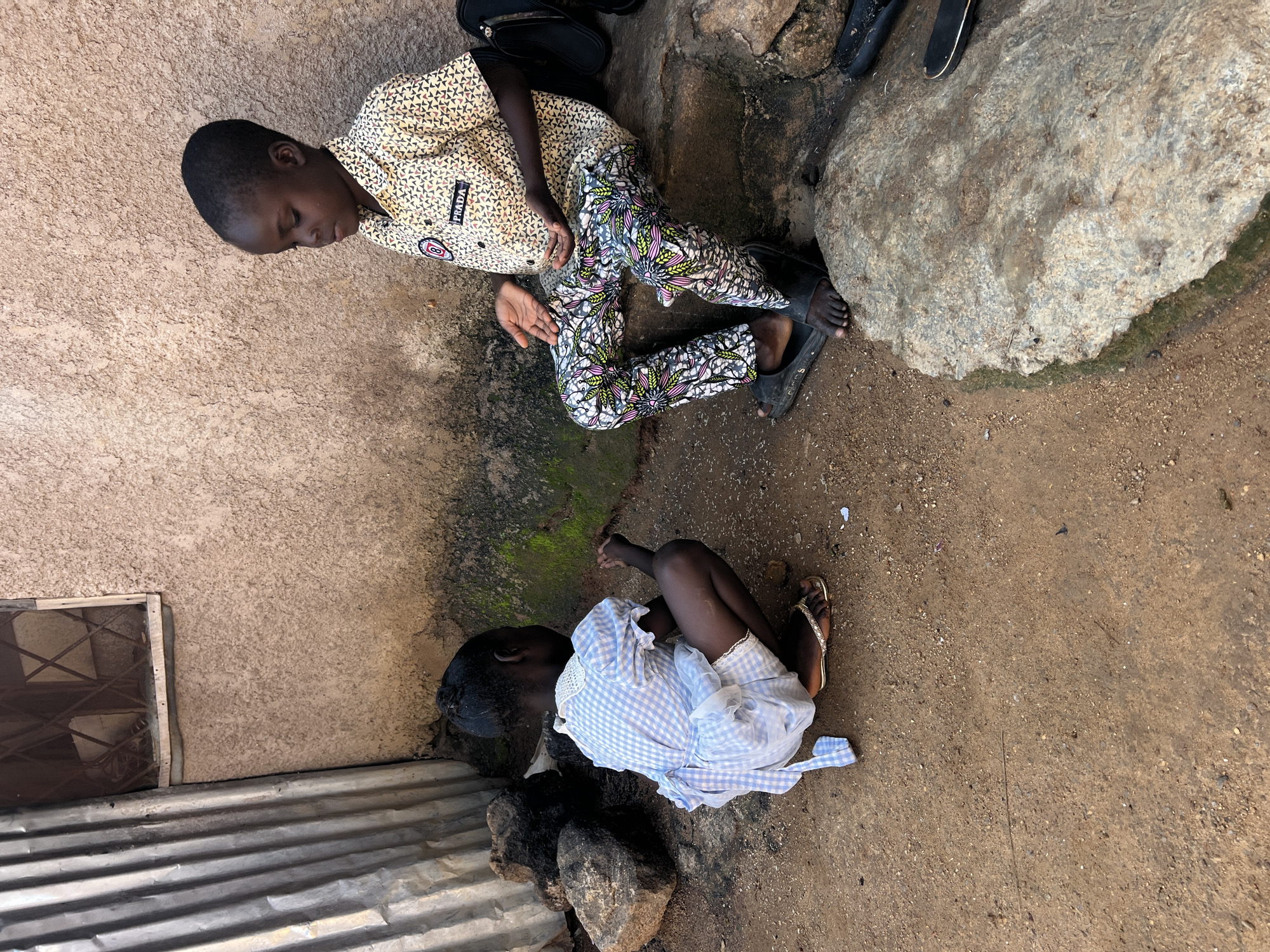 In a dusty, outdoor area in Jos, Nigeria, two young children are interacting on a rough, uneven ground. A boy, seated on a stone ledge, wears a light-colored short-sleeved shirt with a repeating triangular pattern in shades of red, blue, and yellow. His trousers are vividly patterned with large, stylized floral designs in pink, green, and white. On his shirt, a black patch with a white outline and the word "PRADA" in white lettering is visible. He has dark skin and short, dark hair. His hands are open, and he appears to be speaking to the other child.
A younger child, squatting on the ground, is dressed in a light blue and white checkered dress with short, puffed sleeves and a ruffled hem. The dress has a white lace collar. This child also has dark skin and dark hair. They are looking down at their hands, which are clasped together. On their feet, they wear simple, brown flip-flops.
The environment is rustic. To the left of the children is a corrugated metal structure, possibly part of a building or fence, showing vertical ridges. Behind them is a wall made of rough, textured material, appearing to be mud or stucco, with some patches of moss or green growth near the ground. A small, barred window is visible in the upper portion of the wall. Large, dark rocks are scattered around the ground, some appearing to be part of the seating or landscape. The ground itself is a mix of dry dirt, sand, and small pebbles. The overall impression is of a simple, everyday scene in an unpaved area.