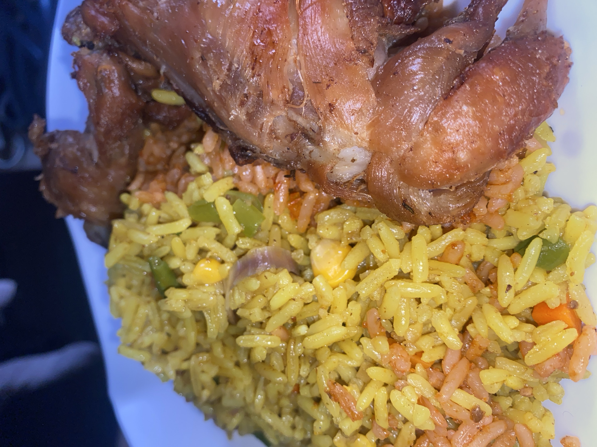 A close-up shot displays a meal served on a white plate, consisting of rice and roasted meat. The rice exhibits a mixed coloration of yellow and reddish-orange grains, intermingled with visible pieces of green bell pepper, corn kernels, and translucent slices of red onion. Adjacent to the rice are multiple pieces of roasted poultry, characterized by their dark brown, crispy skin and visible bone structure, suggesting chicken. The background is dark and out of focus. This scene is situated in the city of Rido, Nigeria.