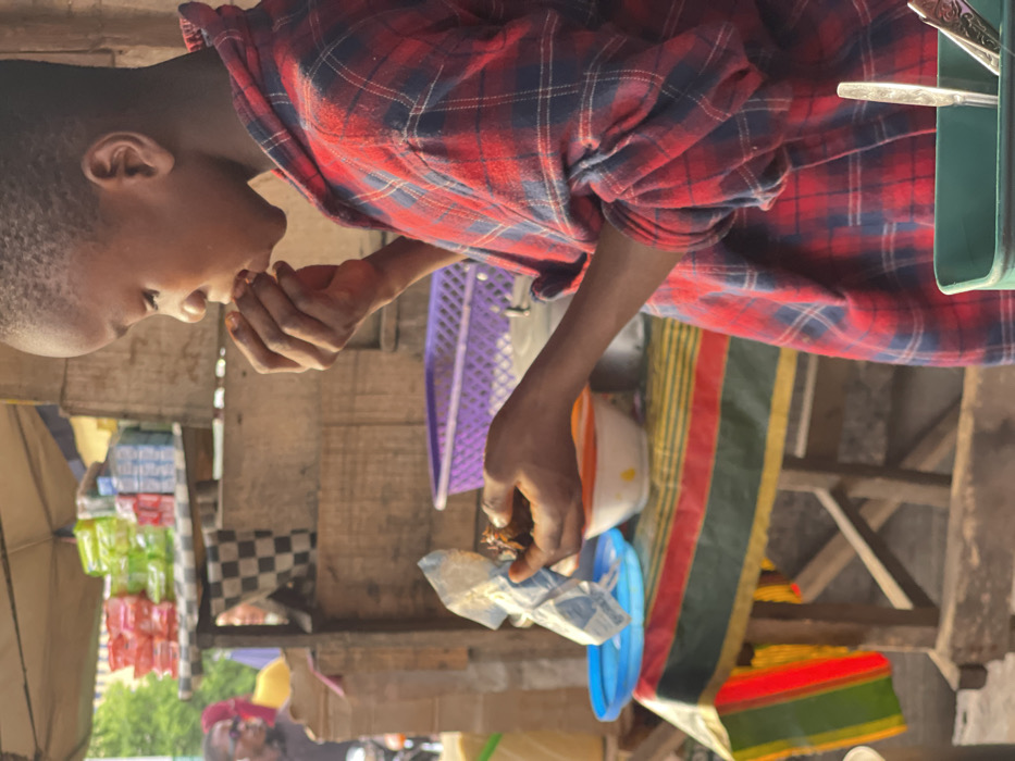 The image, rotated 90 degrees clockwise, shows a young person in profile, likely a boy, eating something with his hands in what appears to be an outdoor market or street stall setting.
The person is positioned in the left half of the frame, facing towards the right. They have short, dark hair and are wearing a red and dark blue plaid long-sleeved shirt. Their right hand is raised to their mouth, and they are in the process of taking a bite of food. Their left hand holds a small, crumpled wrapper or package, from which the food is being consumed.
In the mid-ground, behind the person, a purple plastic crate or basket is visible. To the left and slightly behind the person, various packaged goods in brightly colored wrappers (appearing in shades of blue, green, and red) are stacked or displayed on wooden shelves or a stall. Part of a dark-colored umbrella or canopy is visible on the far left side. The surface beneath these items has a black and white checkered pattern, resembling a checkered cloth or tile.
Further into the mid-ground and extending to the right side of the frame, additional wooden structures suggest market stalls or a framework. On the right side, a colorful striped fabric, featuring prominent green, red, and yellow stripes, is draped over an unseen object. Near this fabric, a white bowl with an orange rim and a blue plastic lid or container are visible. In the upper right corner of the frame, a green container holds several silver utensils, including forks and spoons.
The scene conveys a daytime setting with natural light, suggesting a casual, functional environment, likely a marketplace where goods are sold and food is readily available. The focus is on the person enjoying their meal amidst the backdrop of a bustling, open-air commercial area.