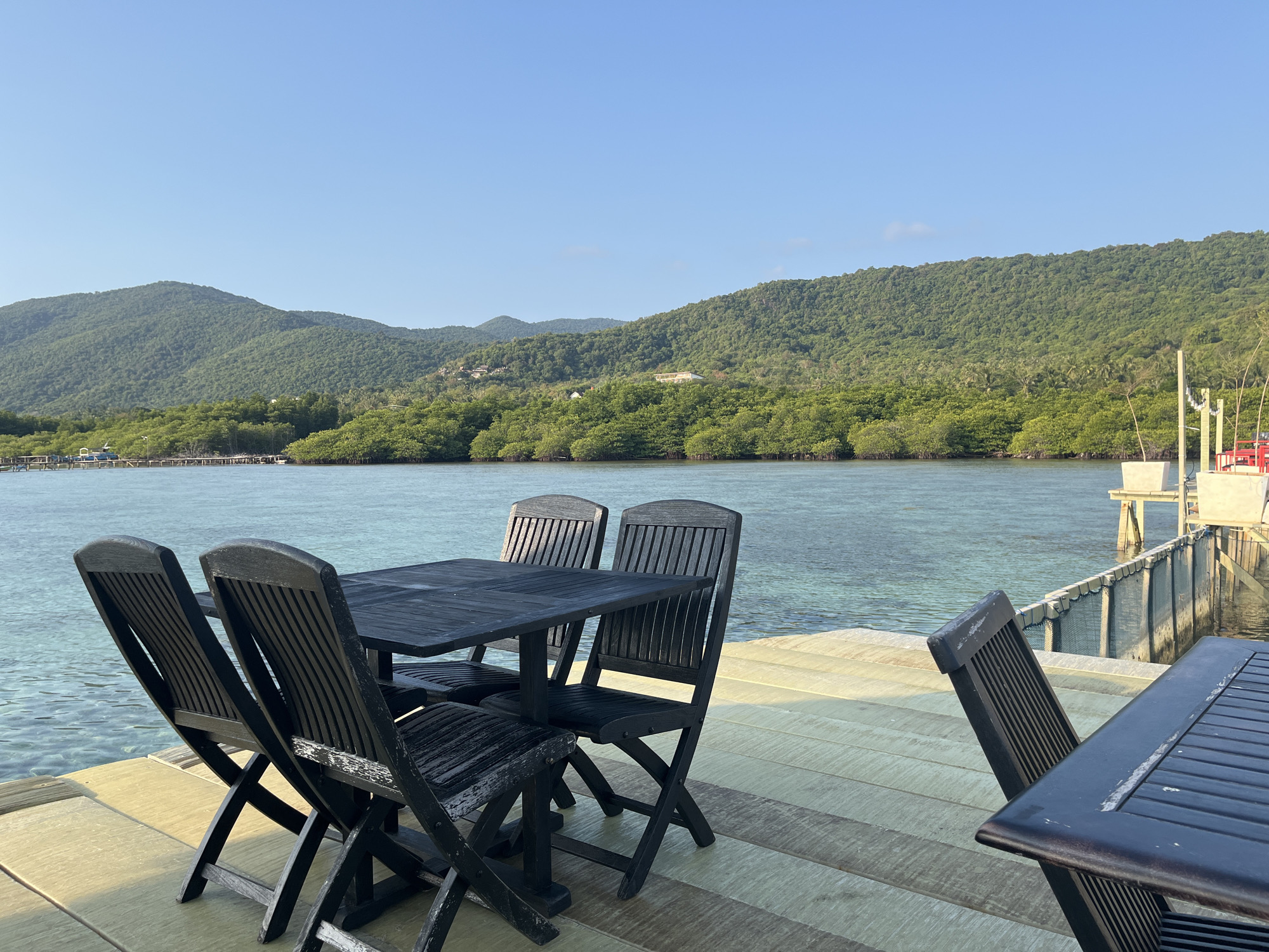 The image depicts an outdoor seating area on a wooden deck overlooking a body of water. A dark-colored rectangular table is surrounded by four matching chairs, all made of a dark wood with slatted designs. The table and chairs appear to be weathered. To the right of the main seating area, another dark table and chair are visible, partially cropped from the frame. The deck itself is made of wooden planks laid horizontally, with some planks appearing lighter in color than others, suggesting wear or different types of wood.
Beyond the deck, a calm body of turquoise water is visible, with gentle ripples on its surface. In the background, a lush, green, tree-covered hillside rises, creating a natural backdrop. The sky above is a clear, pale blue, indicating a sunny day. A wooden pier or walkway extends into the water on the right side of the image, with some structures and fencing visible. The scene suggests a peaceful, scenic location, likely a restaurant or a private deck in a tropical environment. This location is characteristic of the waterfront areas found in the Karimunjawa islands, an archipelago off the coast of Jepara in Central Java, Indonesia, known for its natural beauty and island resorts.