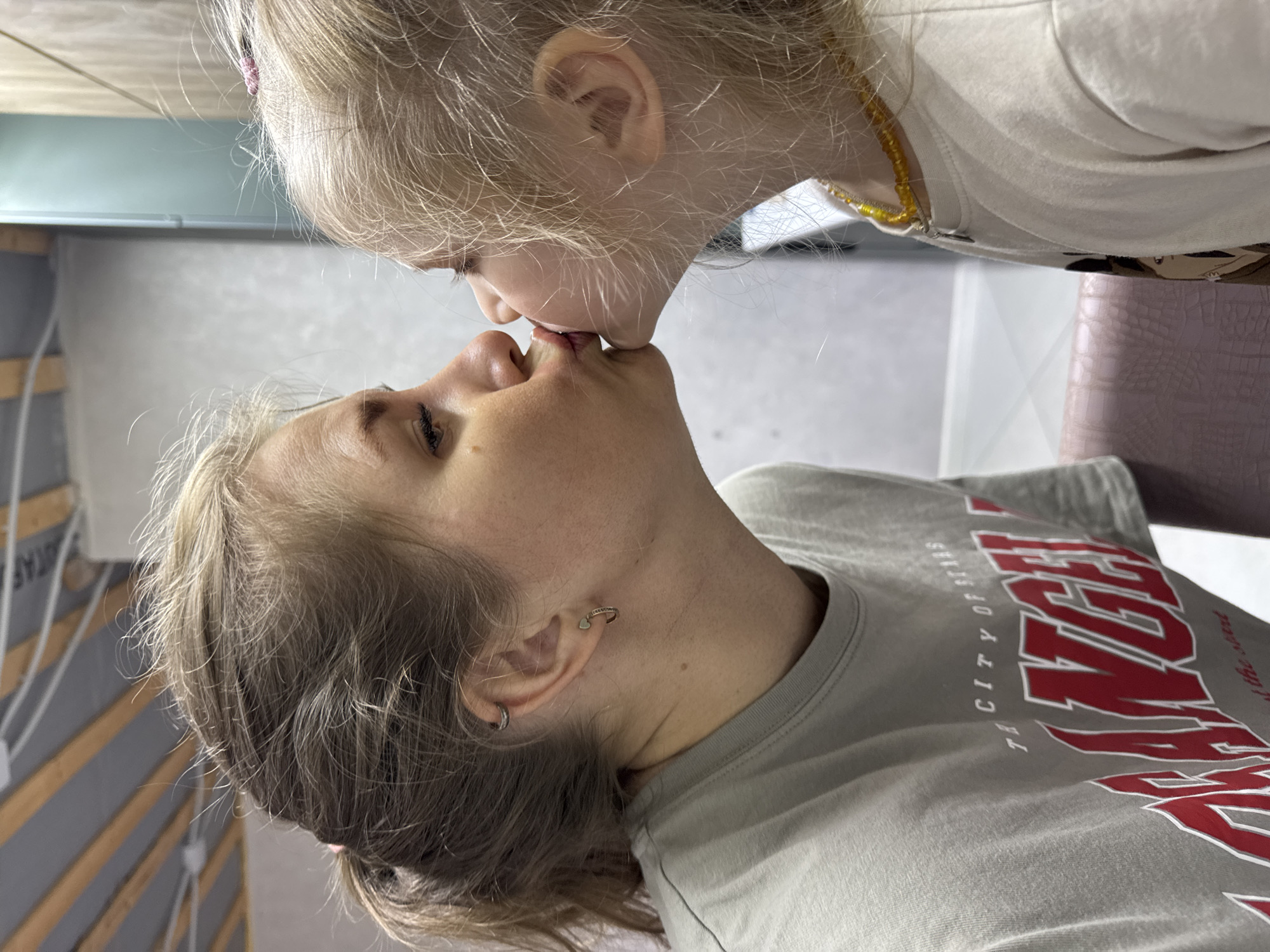 A child kisses an adult on the lips. The child has blonde hair and wears a light-colored shirt. The adult wears a gray T-shirt with the words "LOS ANGELES THE CITY OF STARS" on it and has short blonde hair. The setting appears to be indoors. The location is unknown.